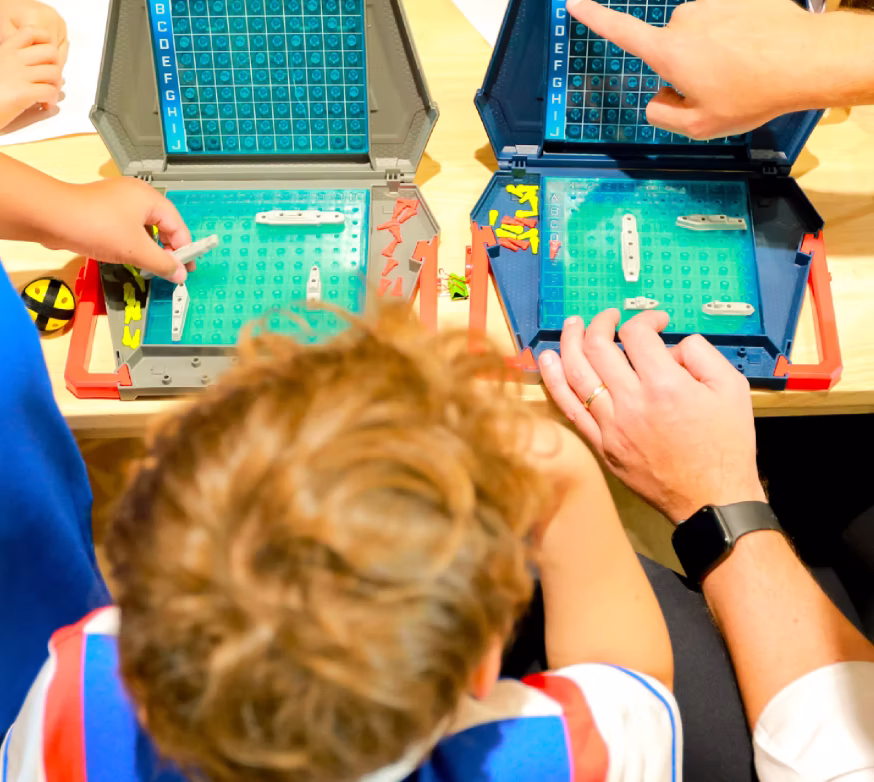 A child and an adult playing Battleship board game, placing ships on blue grids on a table.