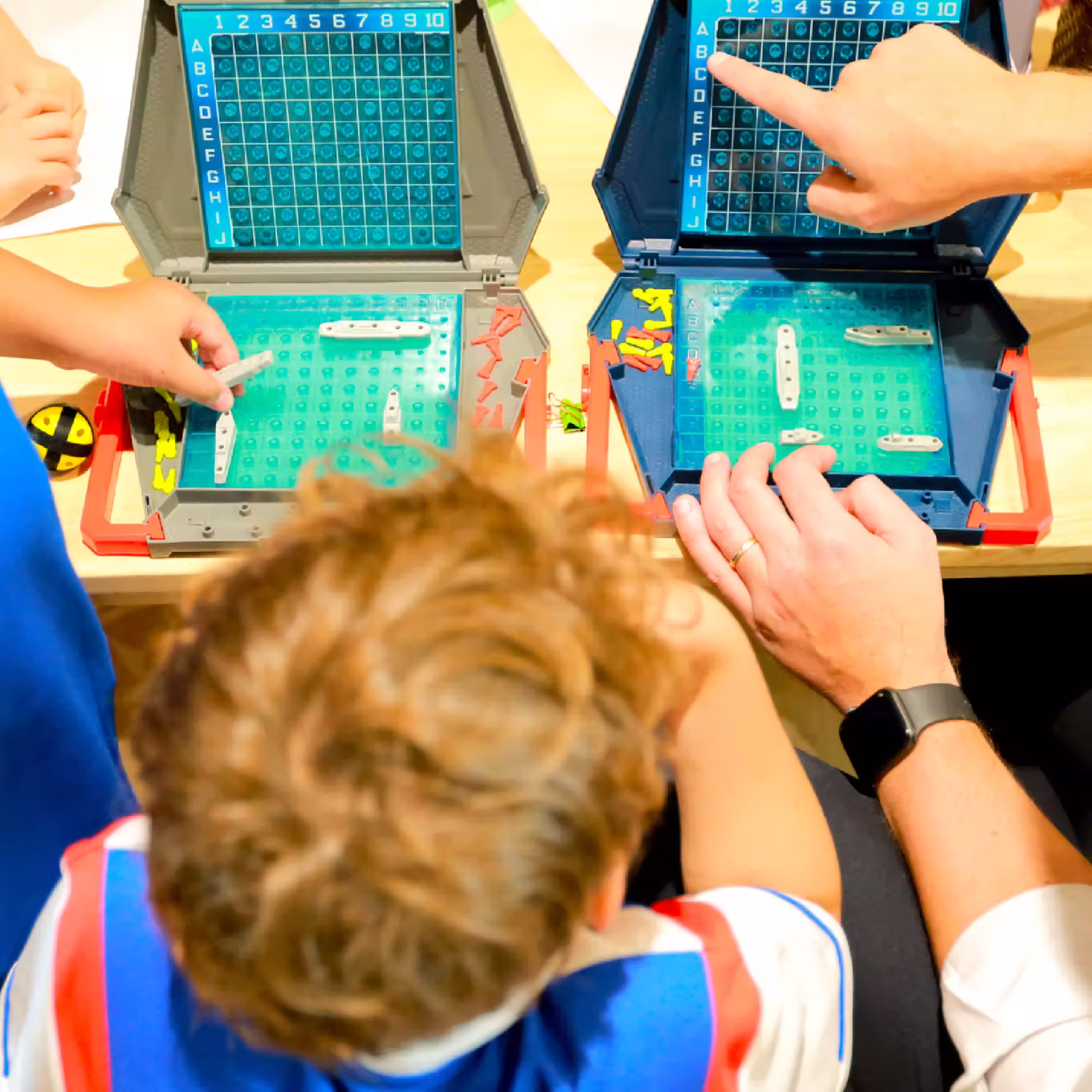 Child and adult playing Battleship board game with plastic grids and ships on a wooden table.