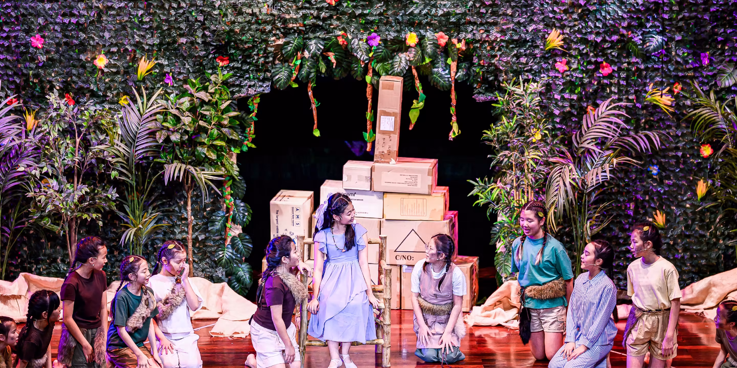 Children in costumes perform a play on stage with a leafy backdrop and stacked boxes as props.