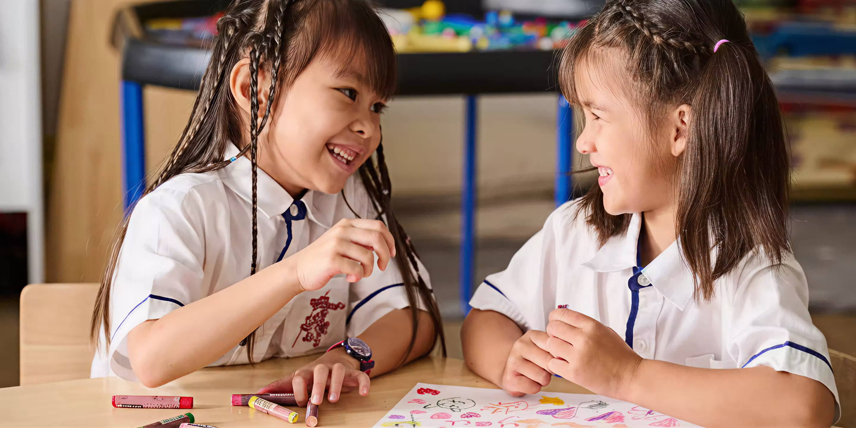 Two young girls in white uniforms laughing and drawing with crayons at a table.
