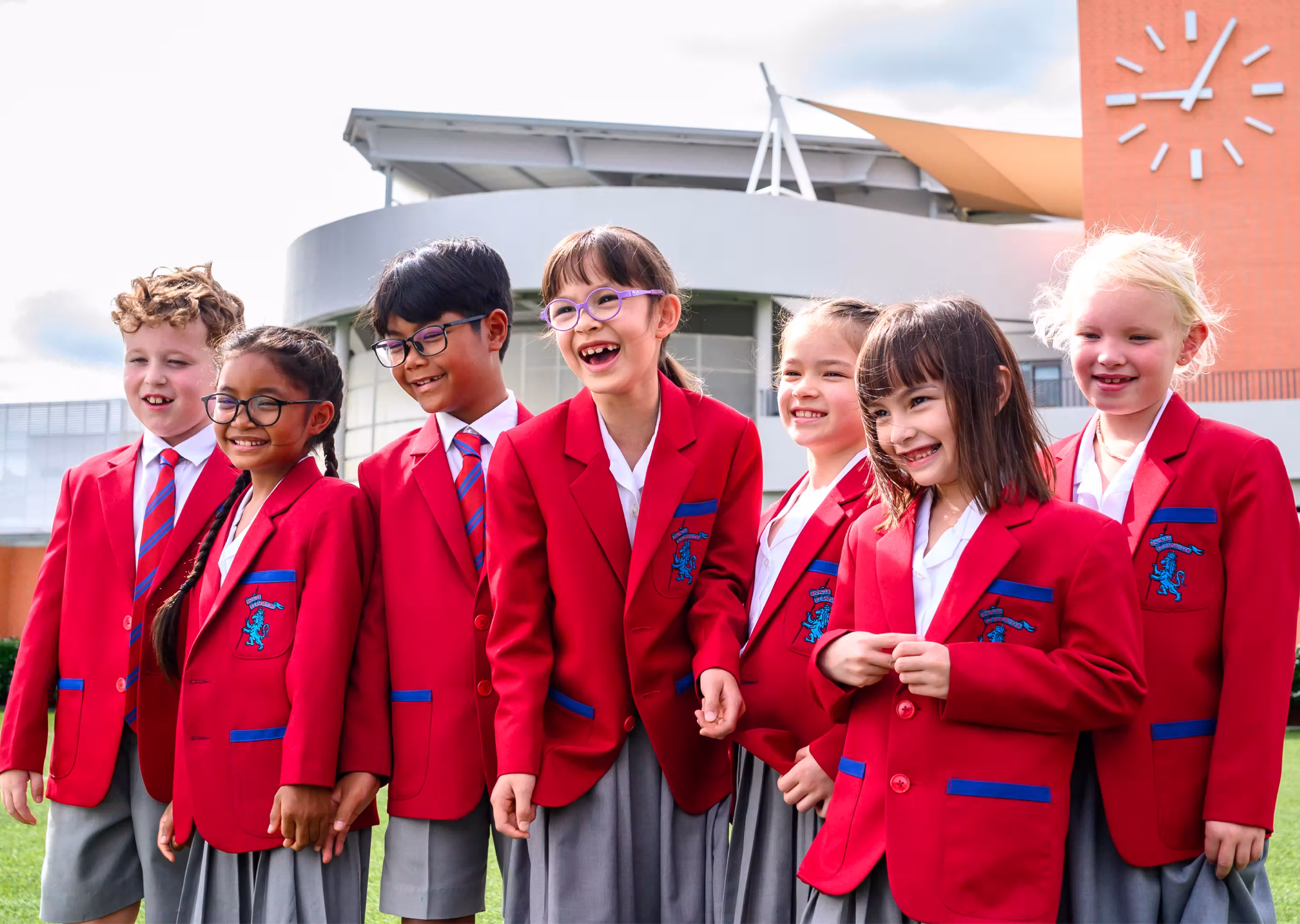 Seven smiling schoolchildren in red blazers and grey skirts standing outside near a clock tower.