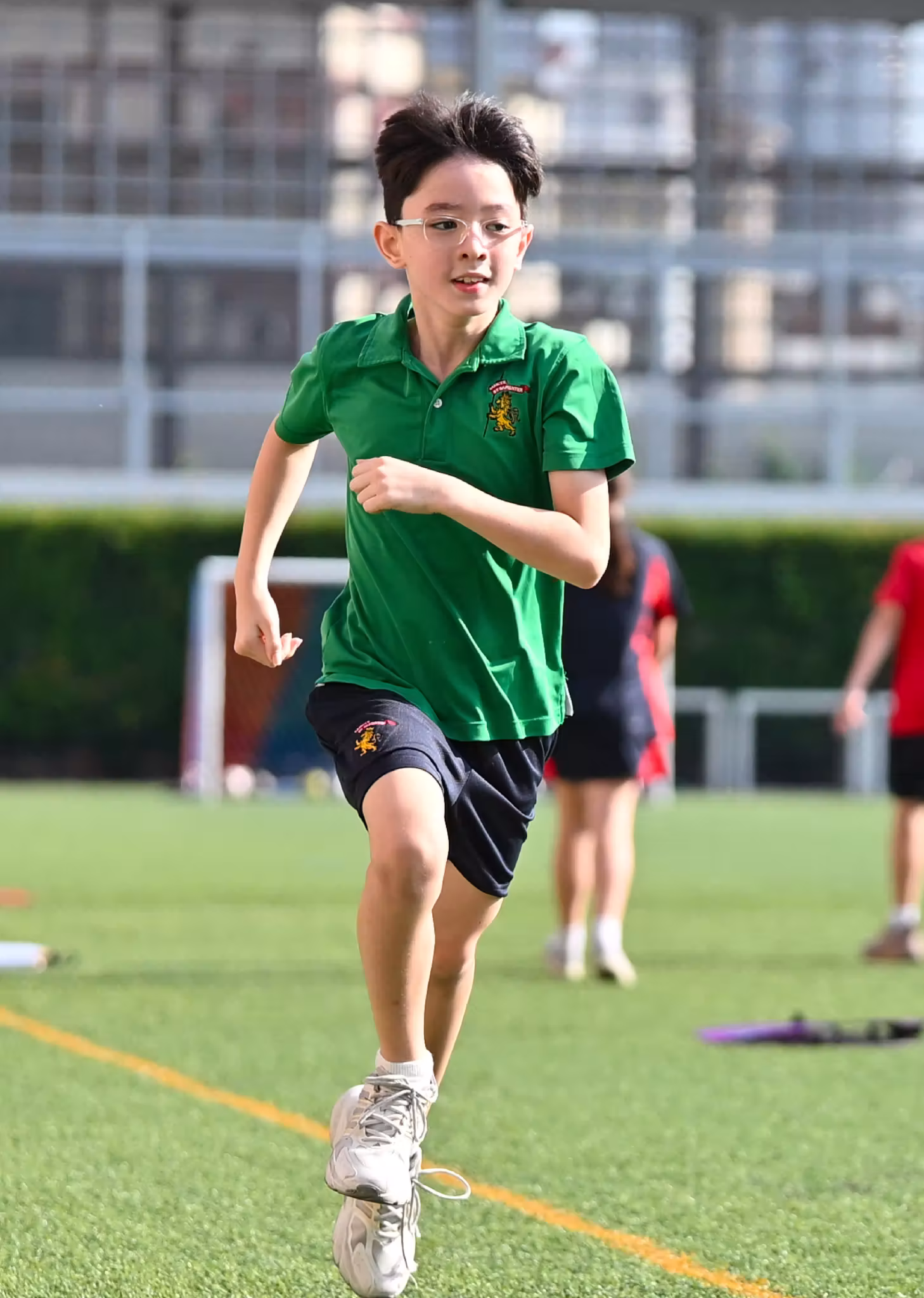 Boy in green shirt and glasses running on a sports field with blurred players in background.