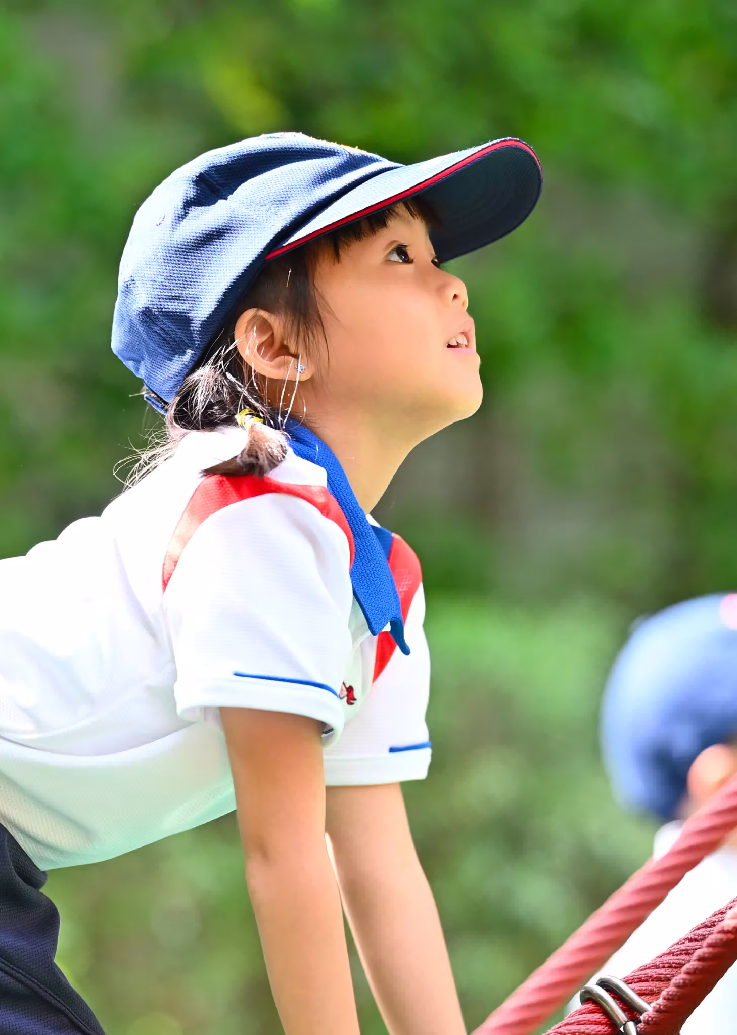 Young girl wearing a blue cap and white shirt looking up while climbing on a playground rope structure.