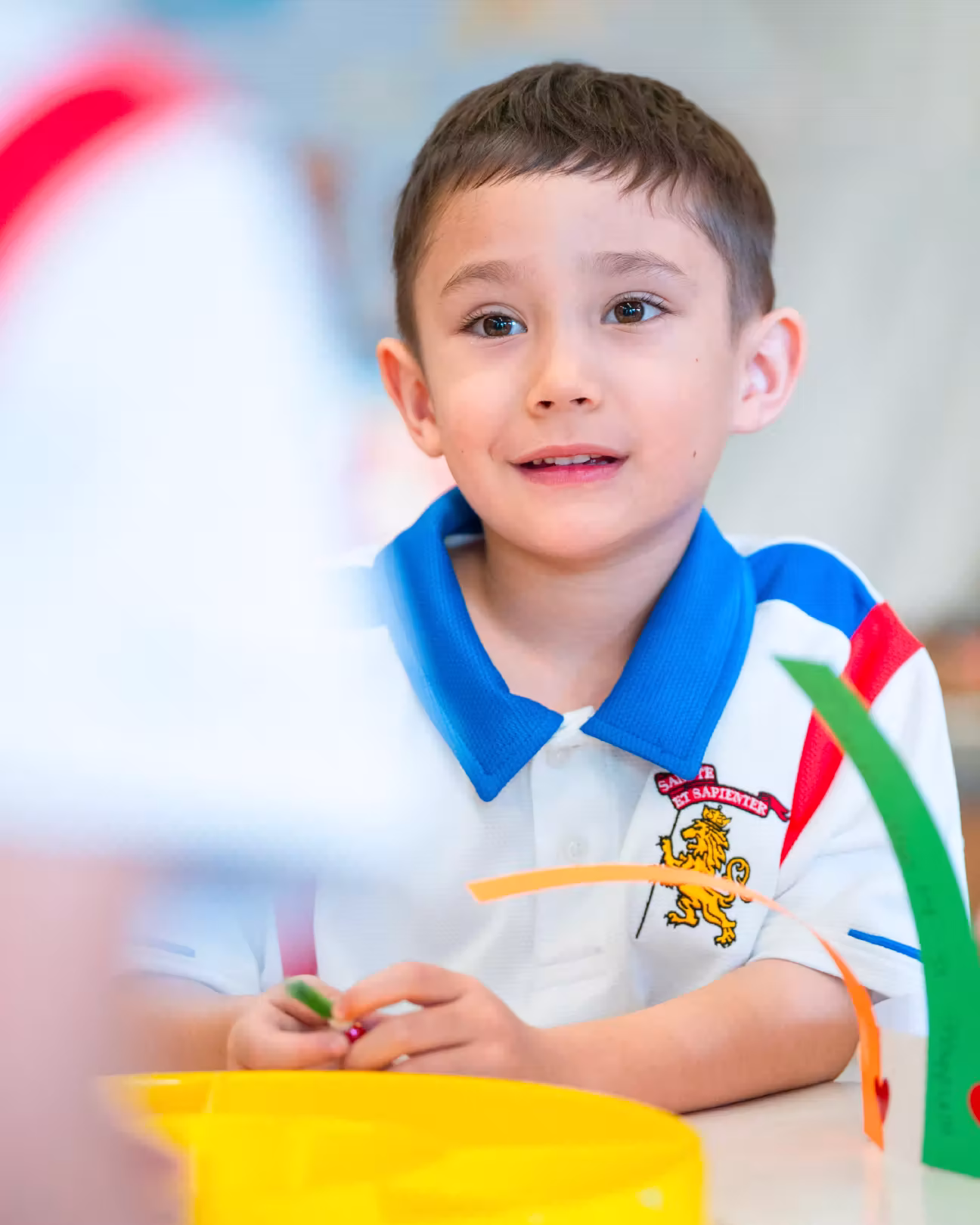 Young boy in a white shirt with blue collar playing with colorful craft strips at a table.