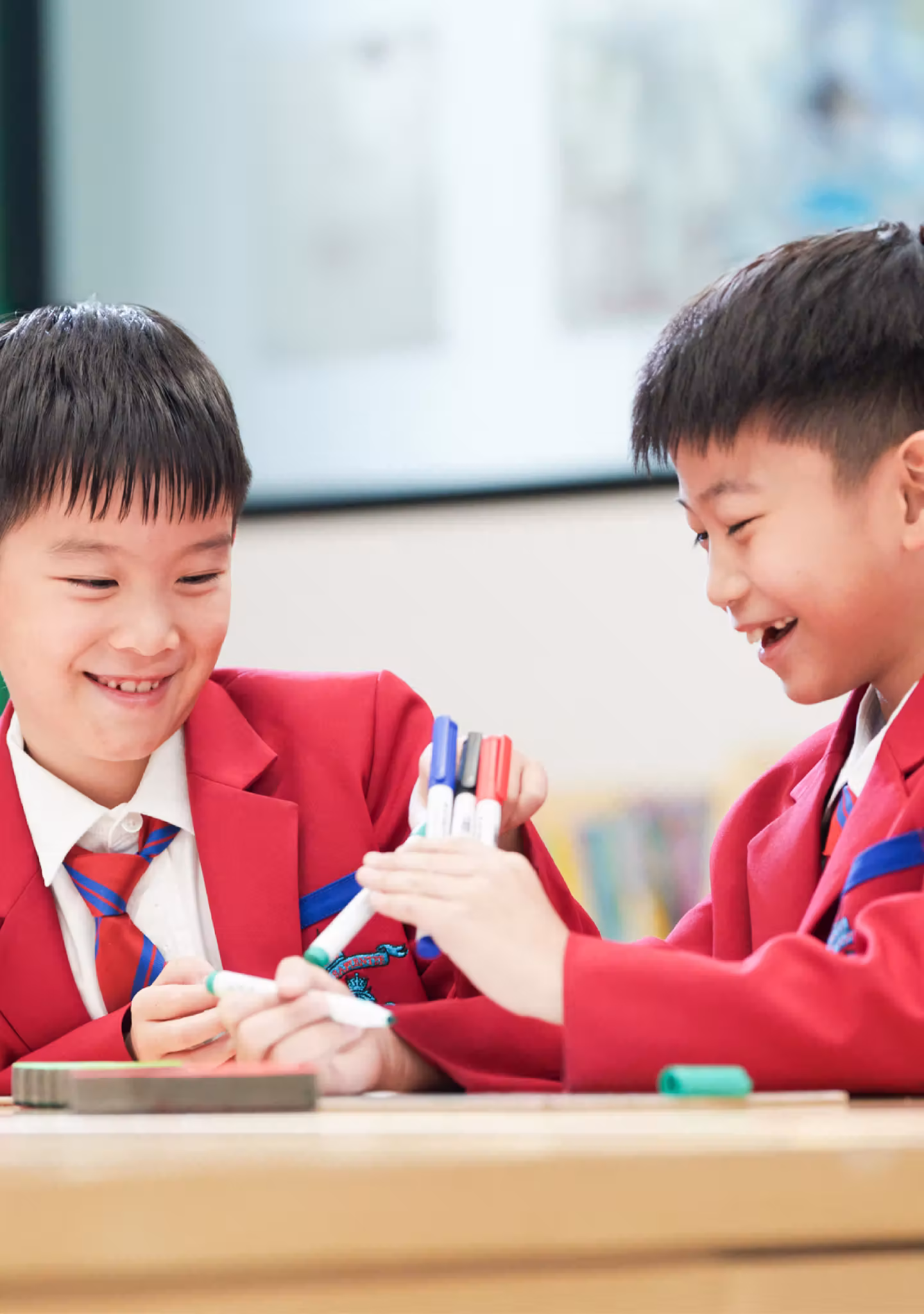 Two boys in red school uniforms smiling and holding colourful markers at a desk in a classroom.