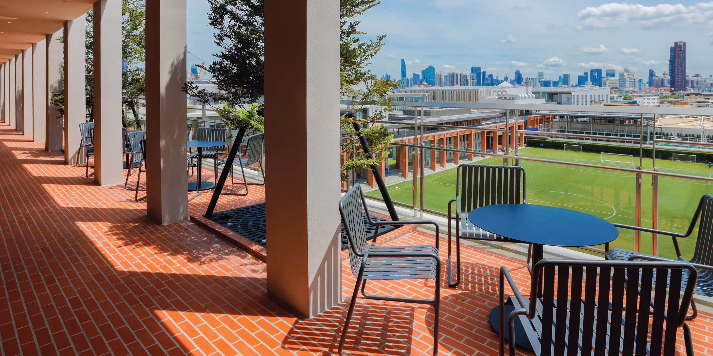 Outdoor seating area with tables and chairs on a terrace overlooking a green sports field and city skyline.
