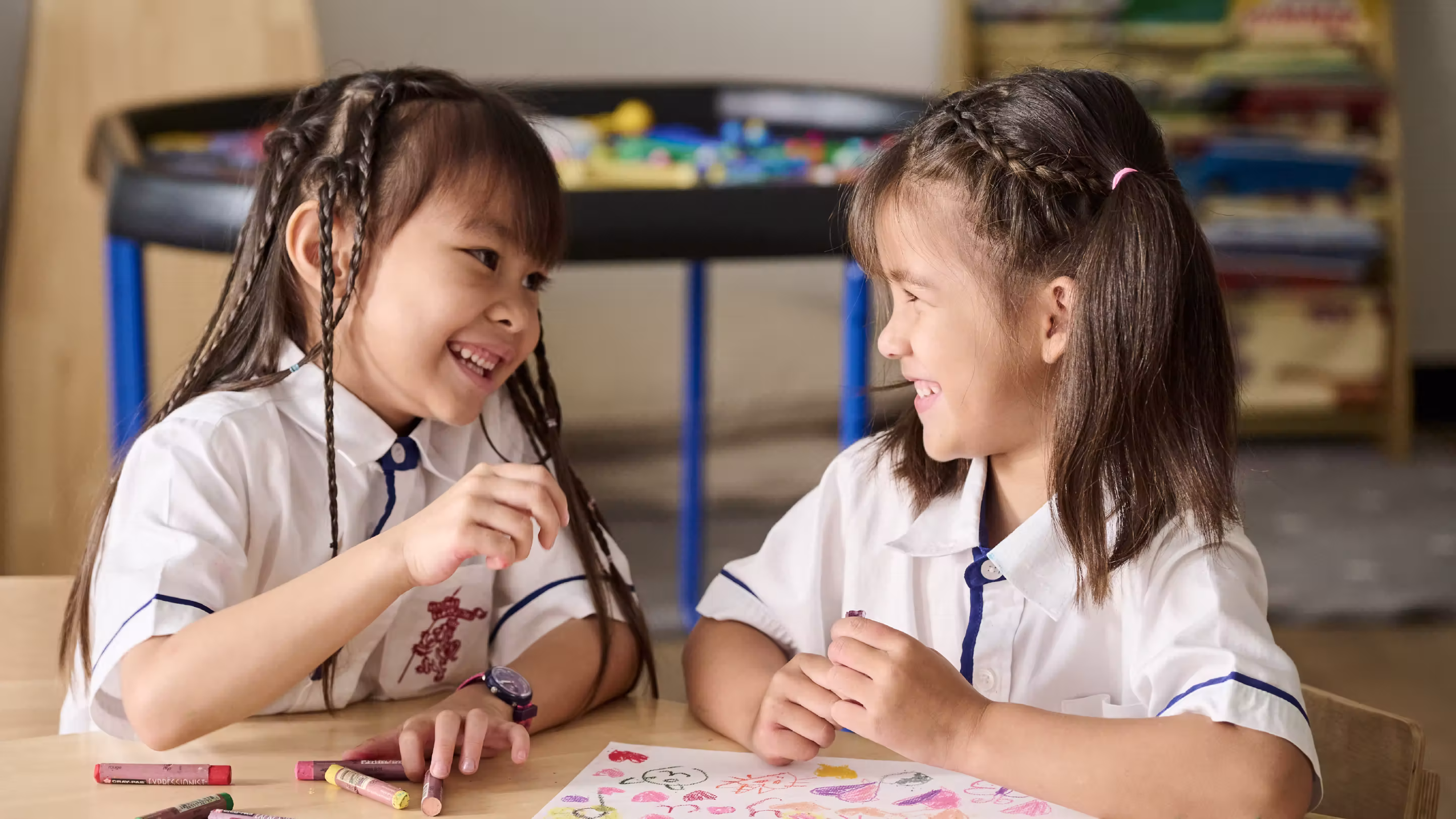 Two young girls in white uniforms smiling and drawing with crayons at a table.