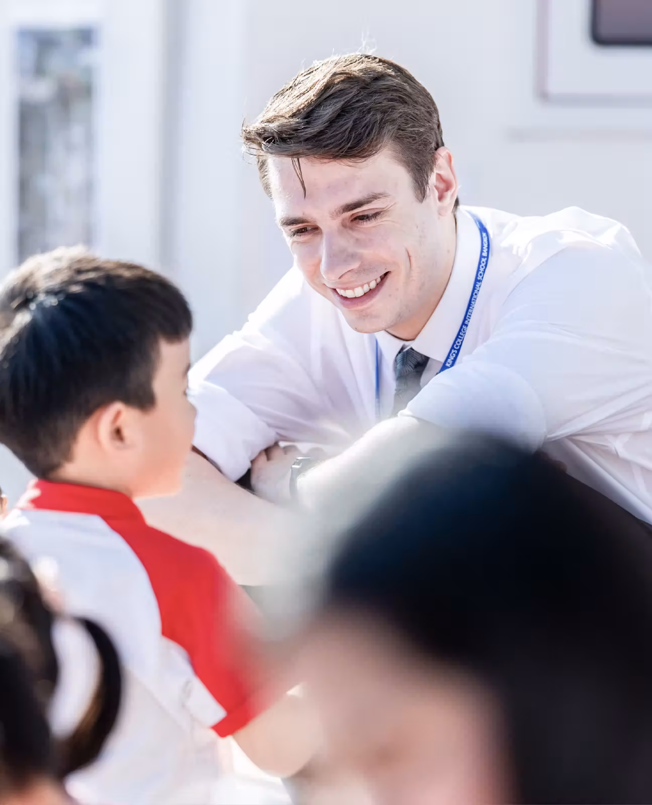 Smiling teacher wearing a lanyard leans forward to talk with a young student in a red and white shirt.