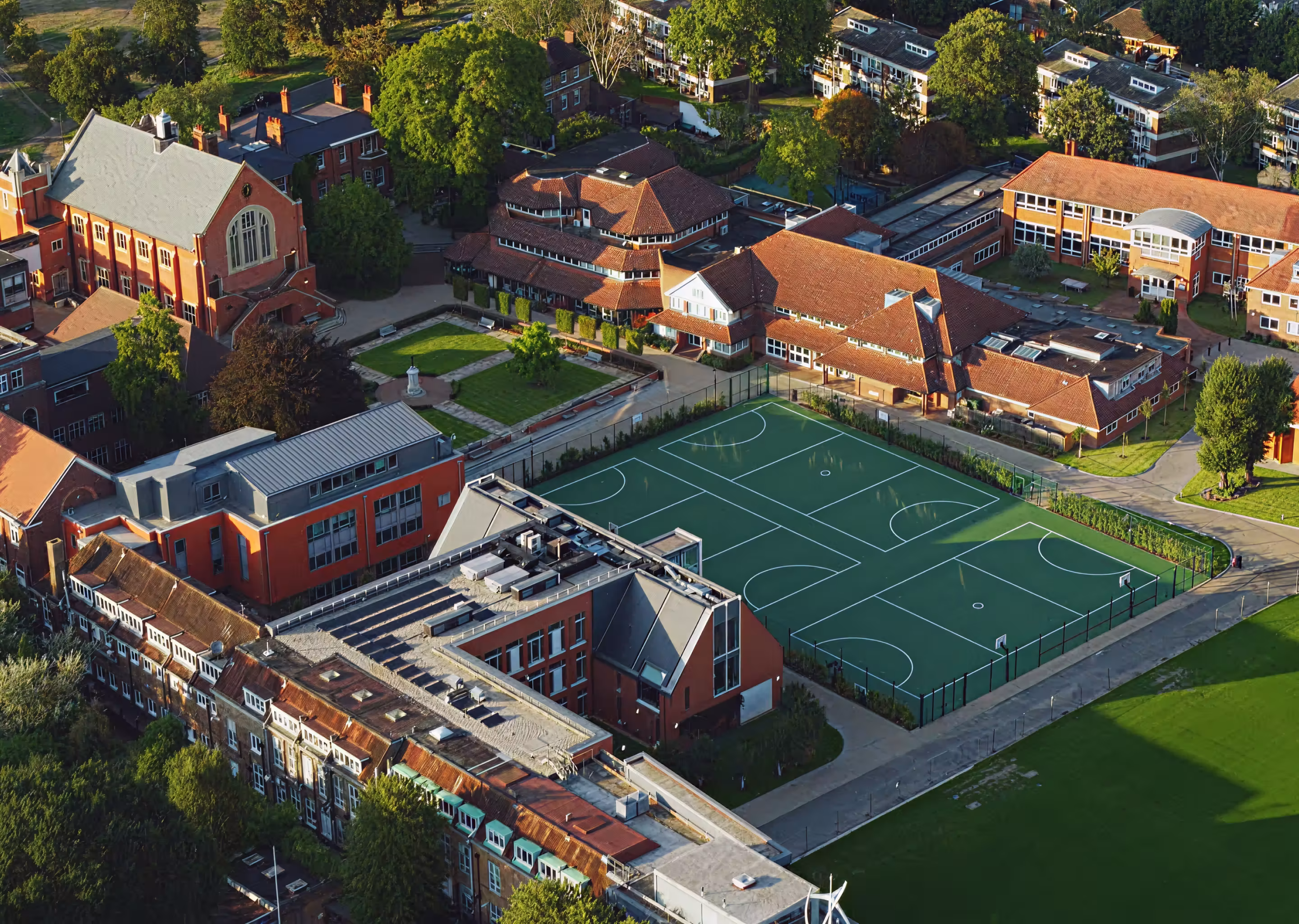 Aerial view of school buildings with a fenced sports court and green playing fields surrounded by trees.
