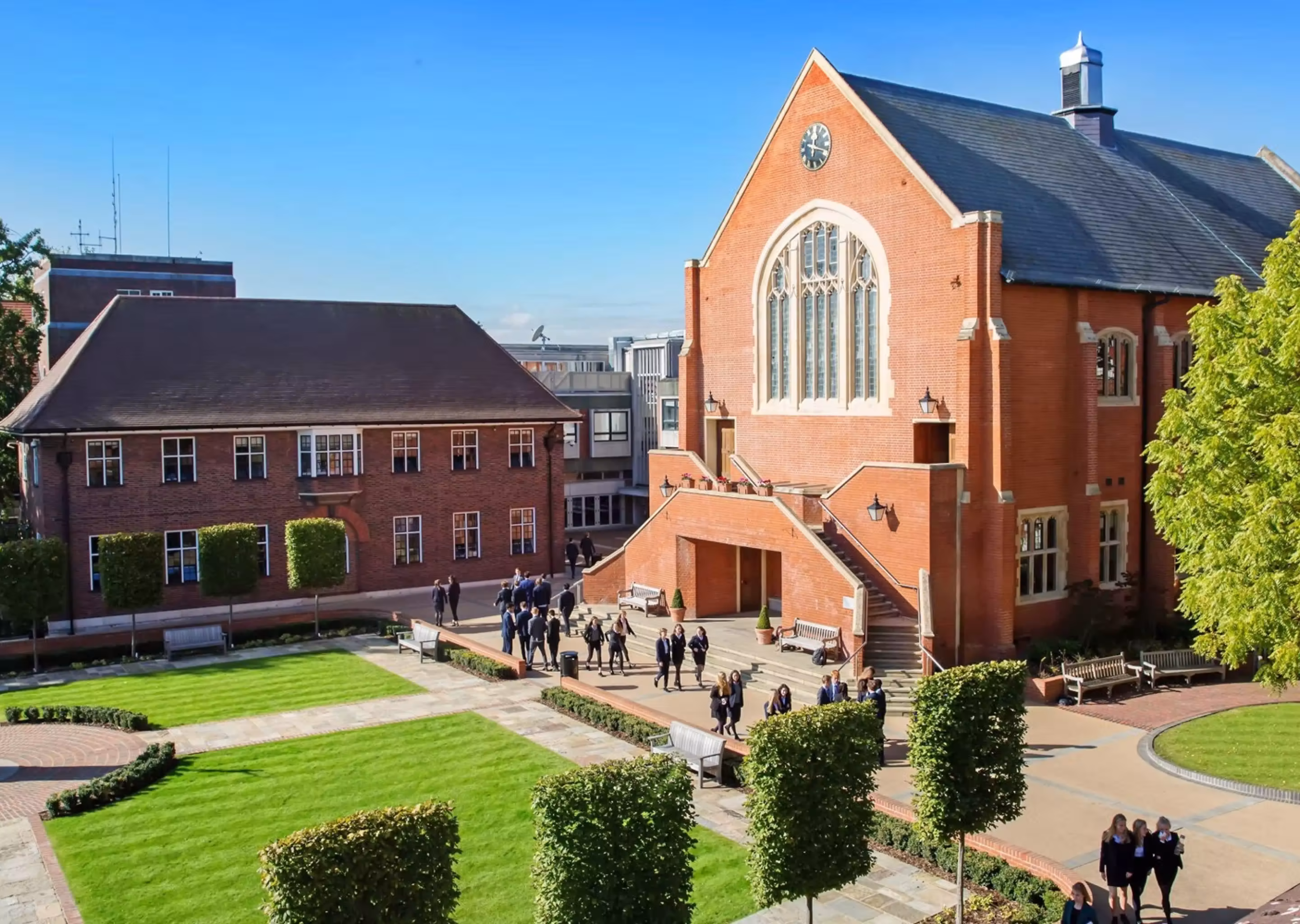 Students walking on paved paths near red brick buildings and trimmed lawns under blue sky.