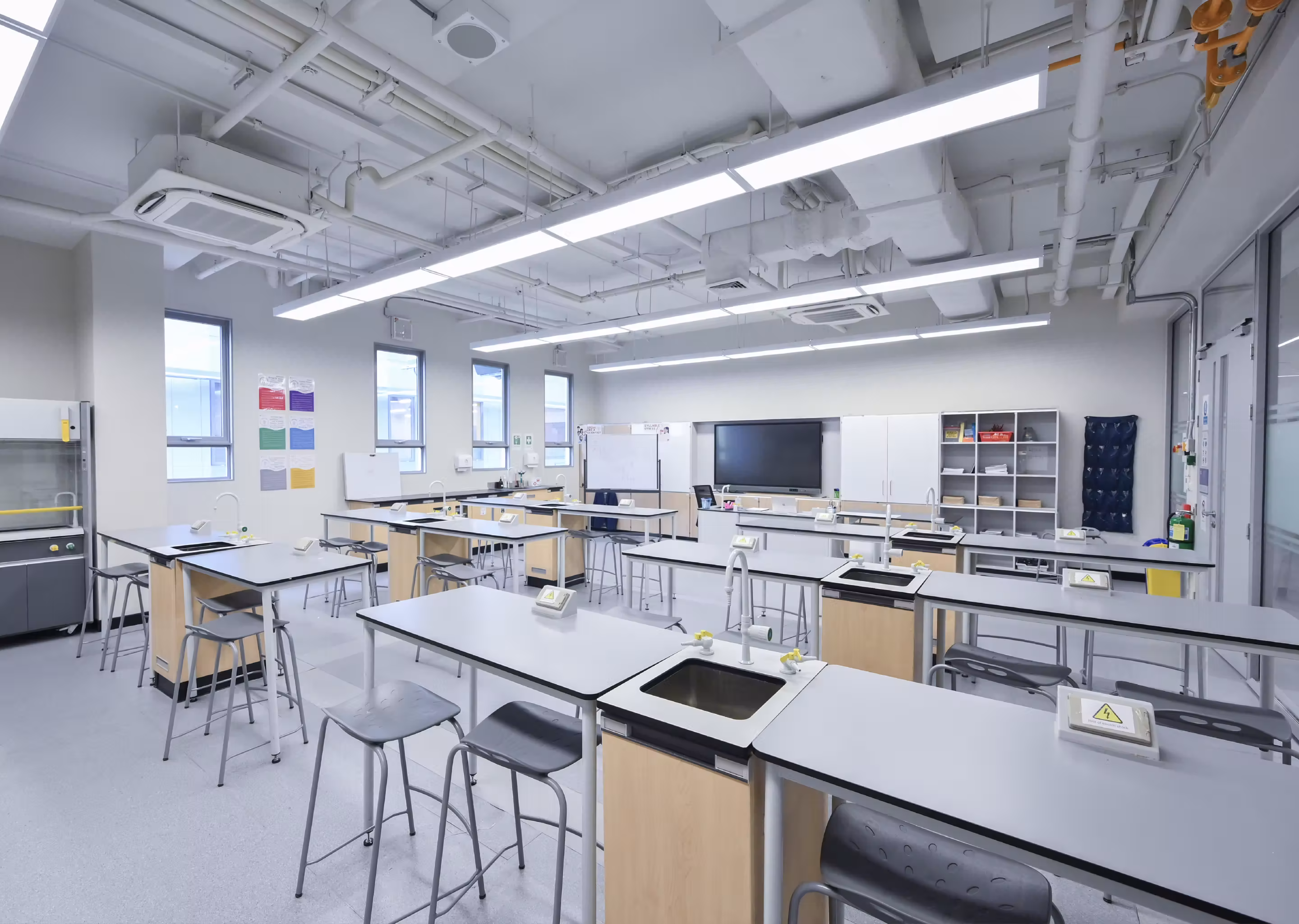 Bright, clean science classroom with lab tables, sinks, stools, and a large screen at the front.
