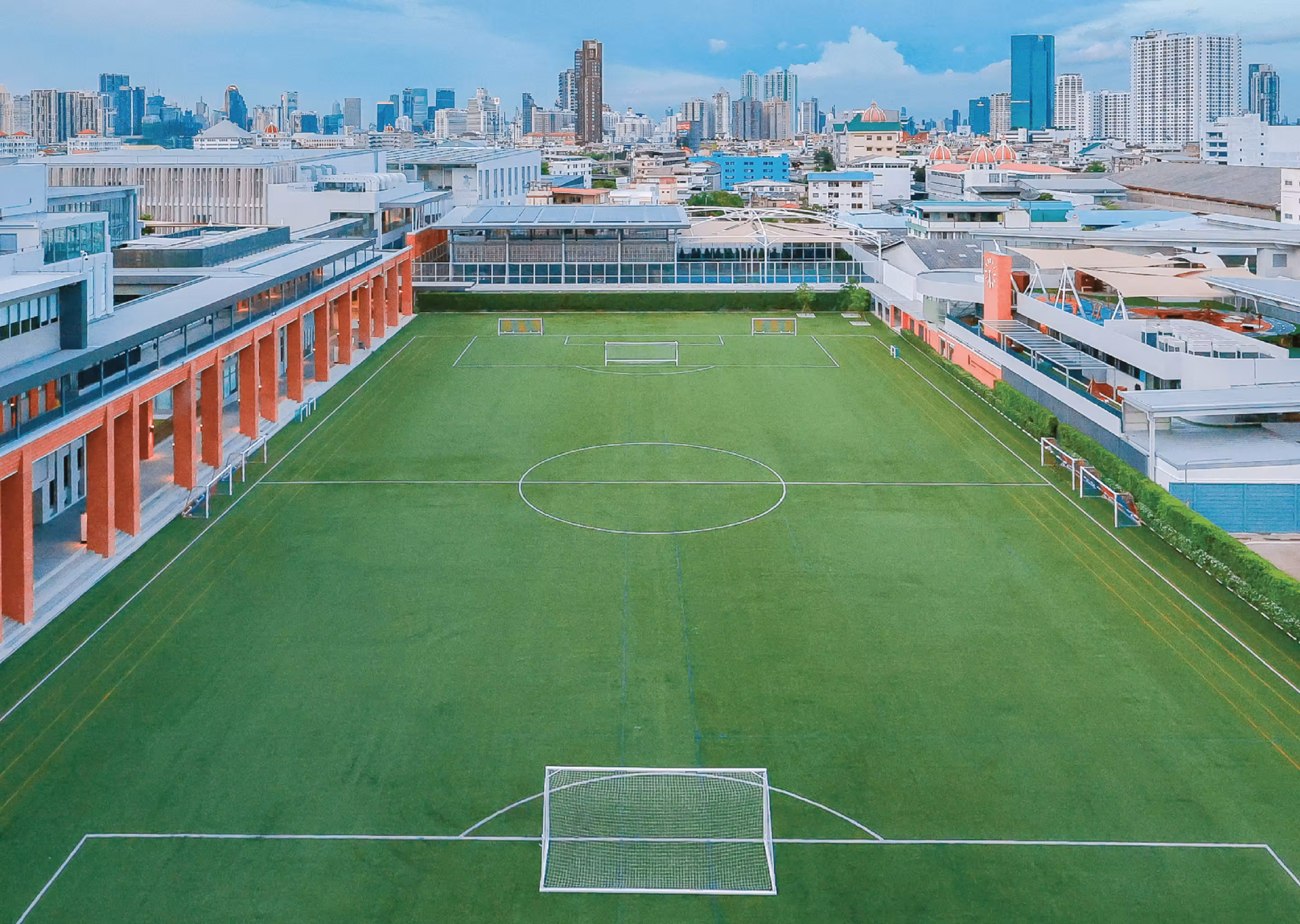 Aerial view of a green football pitch with multiple goals surrounded by buildings and city skyline.
