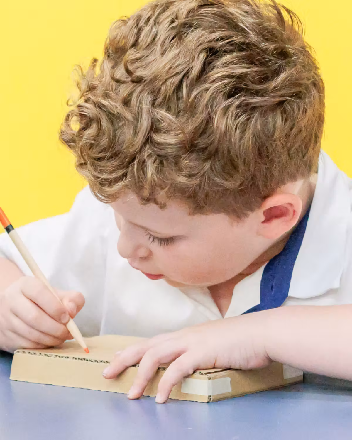 Young boy with curly hair drawing on a cardboard piece with a crayon at a table.