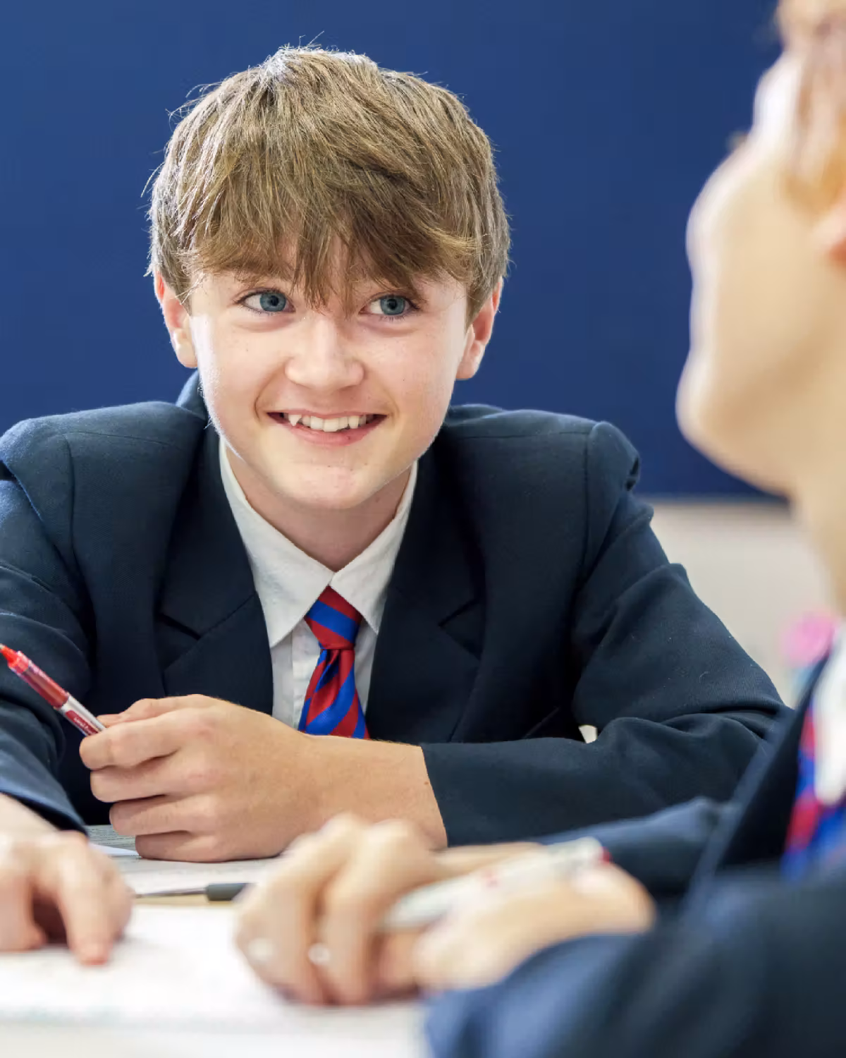 Smiling schoolboy in blazer and striped tie holding a pen, engaging in classroom discussion.