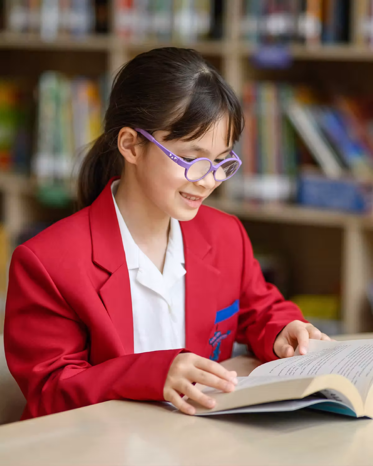Girl in red blazer and purple glasses reading a book at a table in a library.