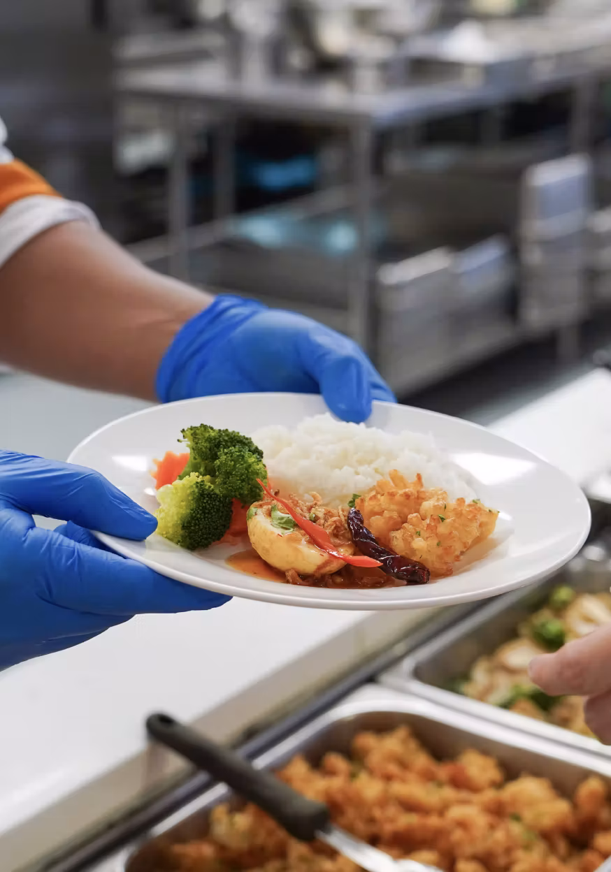 Person wearing blue gloves serving a plate with broccoli, rice, fried food, and chili in a cafeteria.