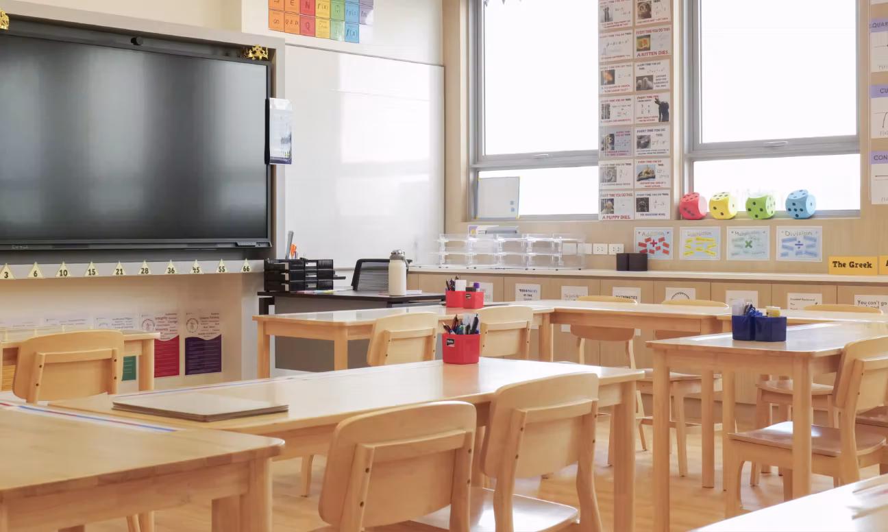 Bright classroom with wooden desks, chairs, large screen, educational posters, and colourful dice on window sill.