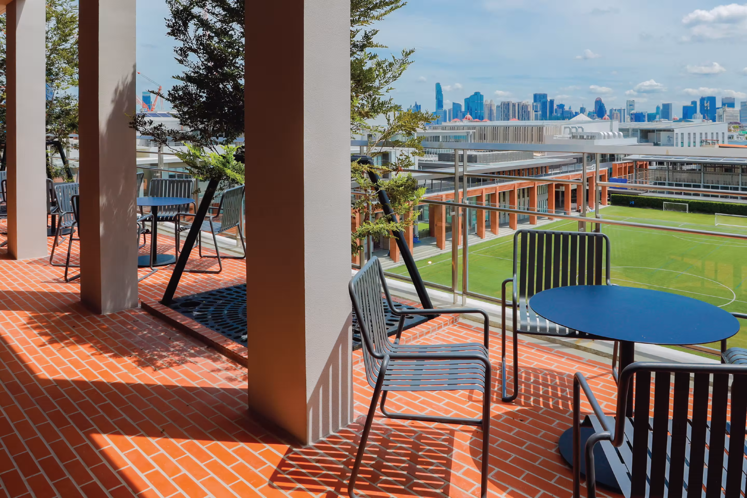 Outdoor terrace with blue tables and chairs overlooking a green sports field and city skyline.