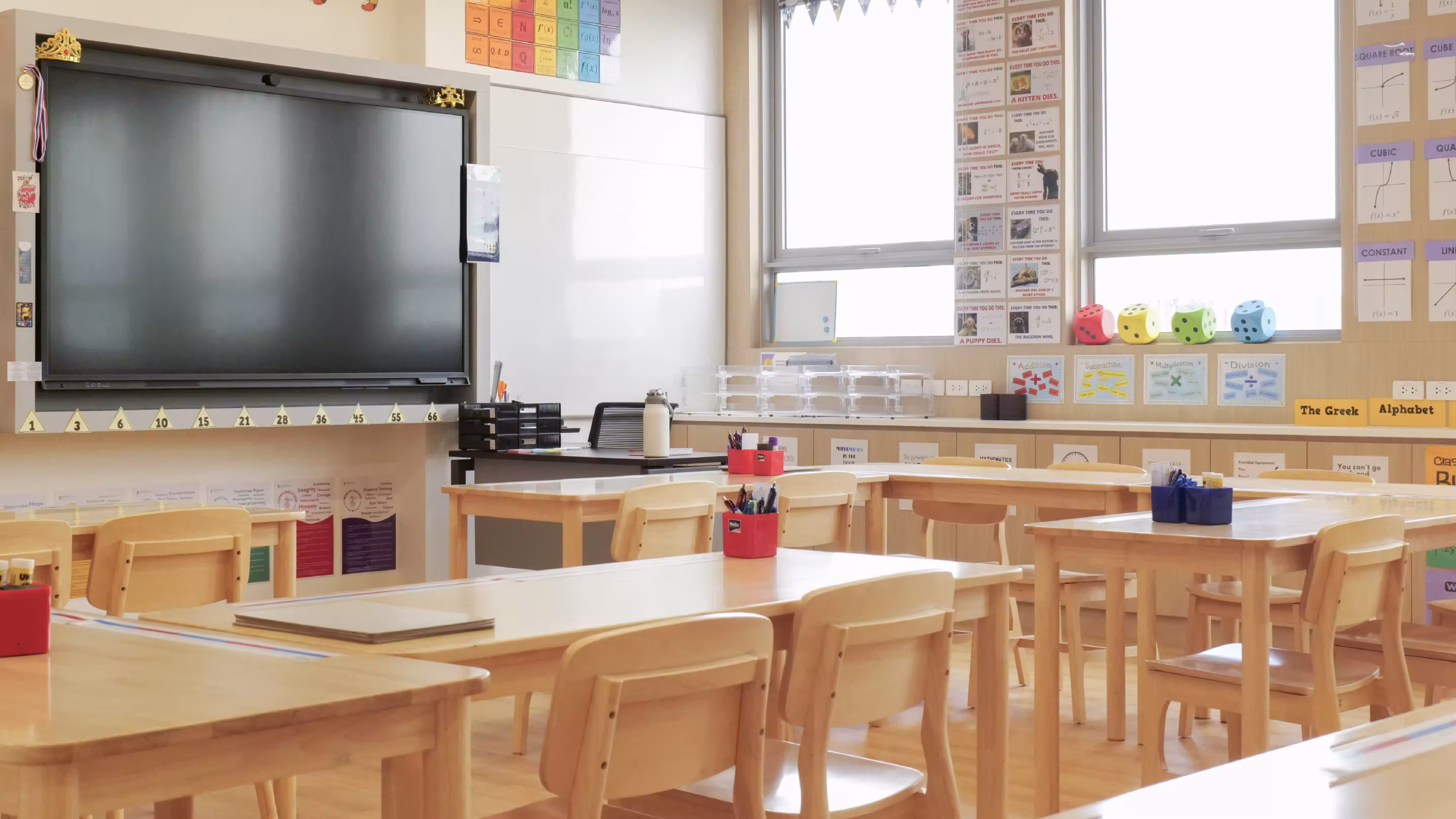 Bright classroom with wooden desks, chairs, a large screen, and educational posters on walls.