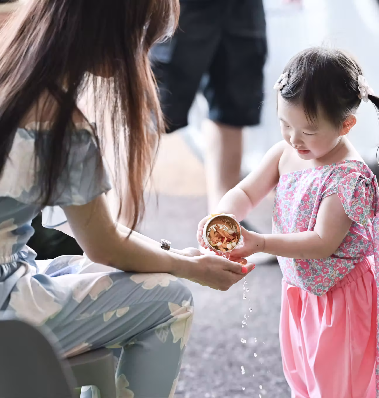 Young girl pours water and flower petals from a cup into a seated woman's cupped hands.