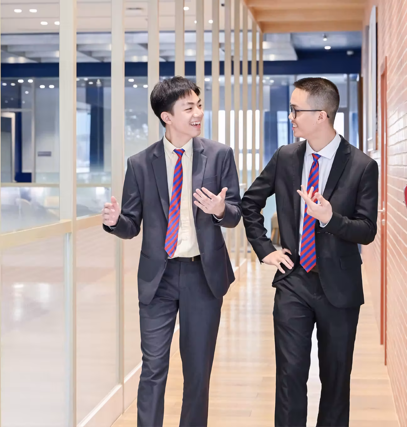 Two young men in suits with blue and red striped ties walking and chatting in a modern office hallway.