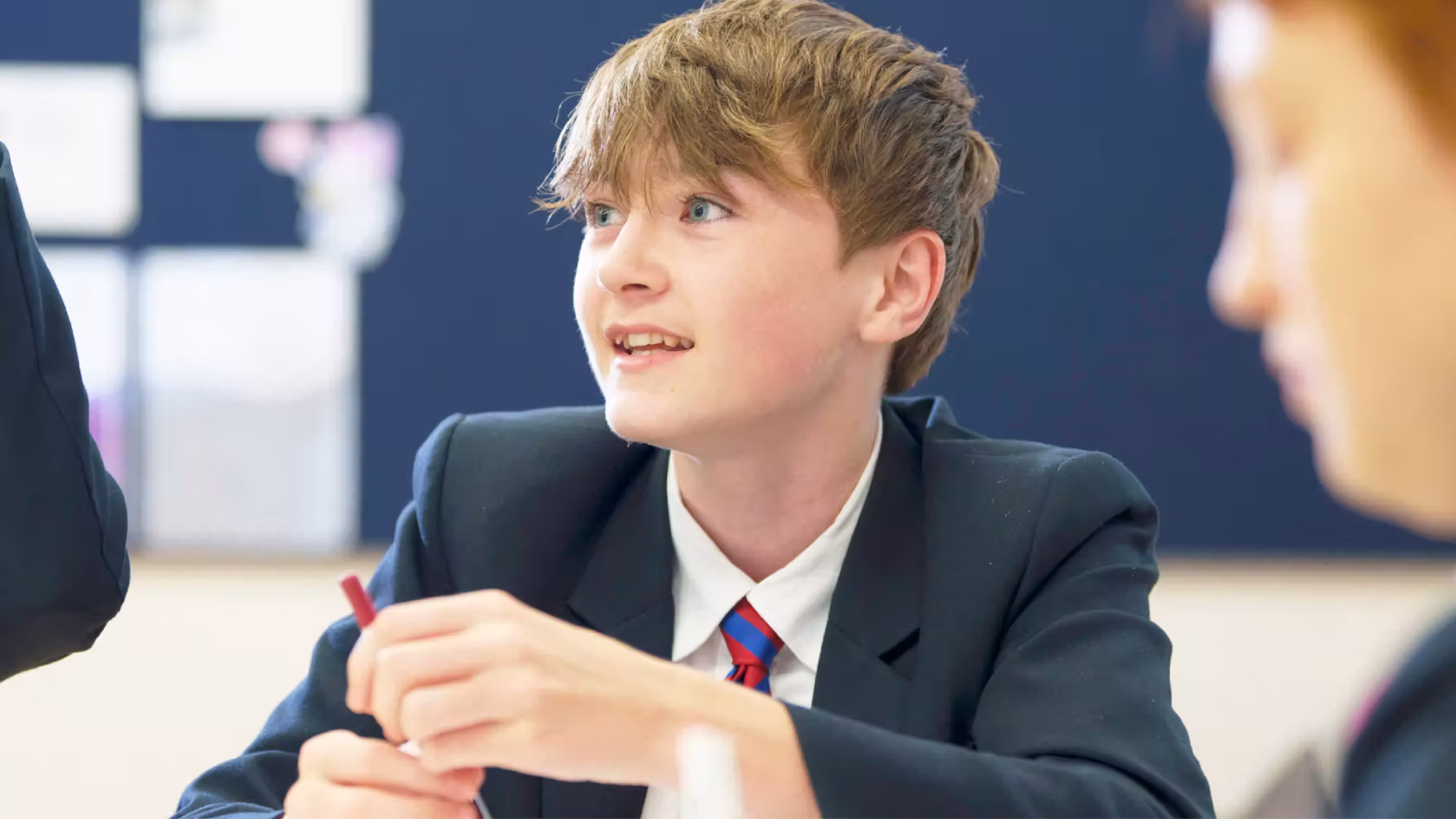 Schoolboy in uniform holding a pencil, looking attentively to his left in a classroom setting.