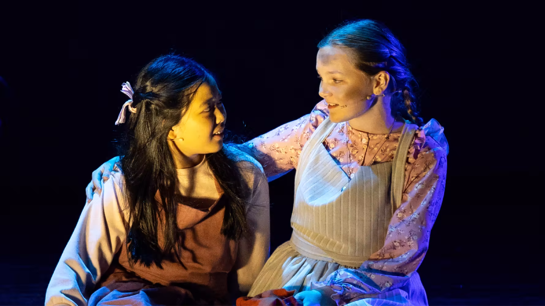 Two young women in period costumes seated, one with arm around the other, looking at each other on stage.
