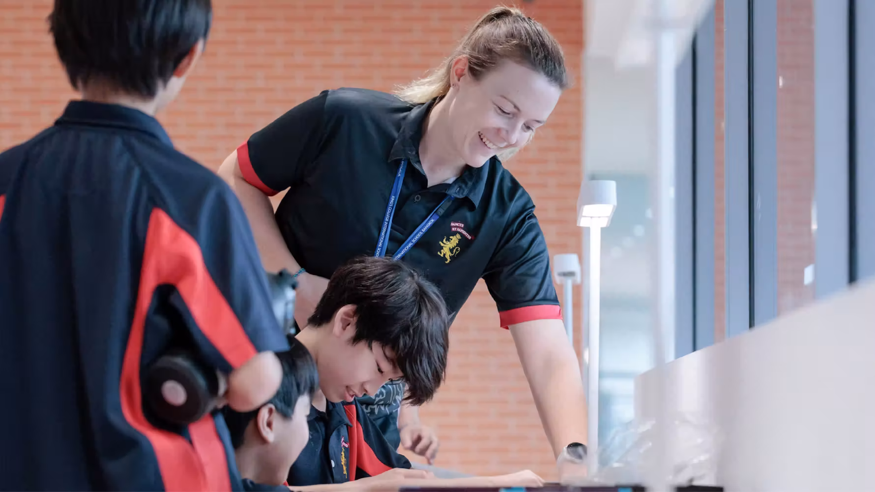 Teacher smiling and engaging with two students in black and red school uniforms near a window.