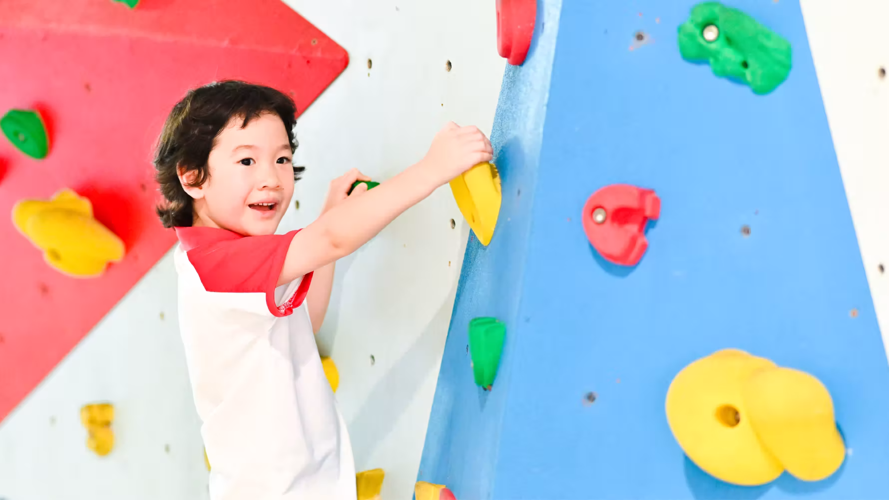 Young boy climbing an indoor rock wall with colorful handholds in a play area.