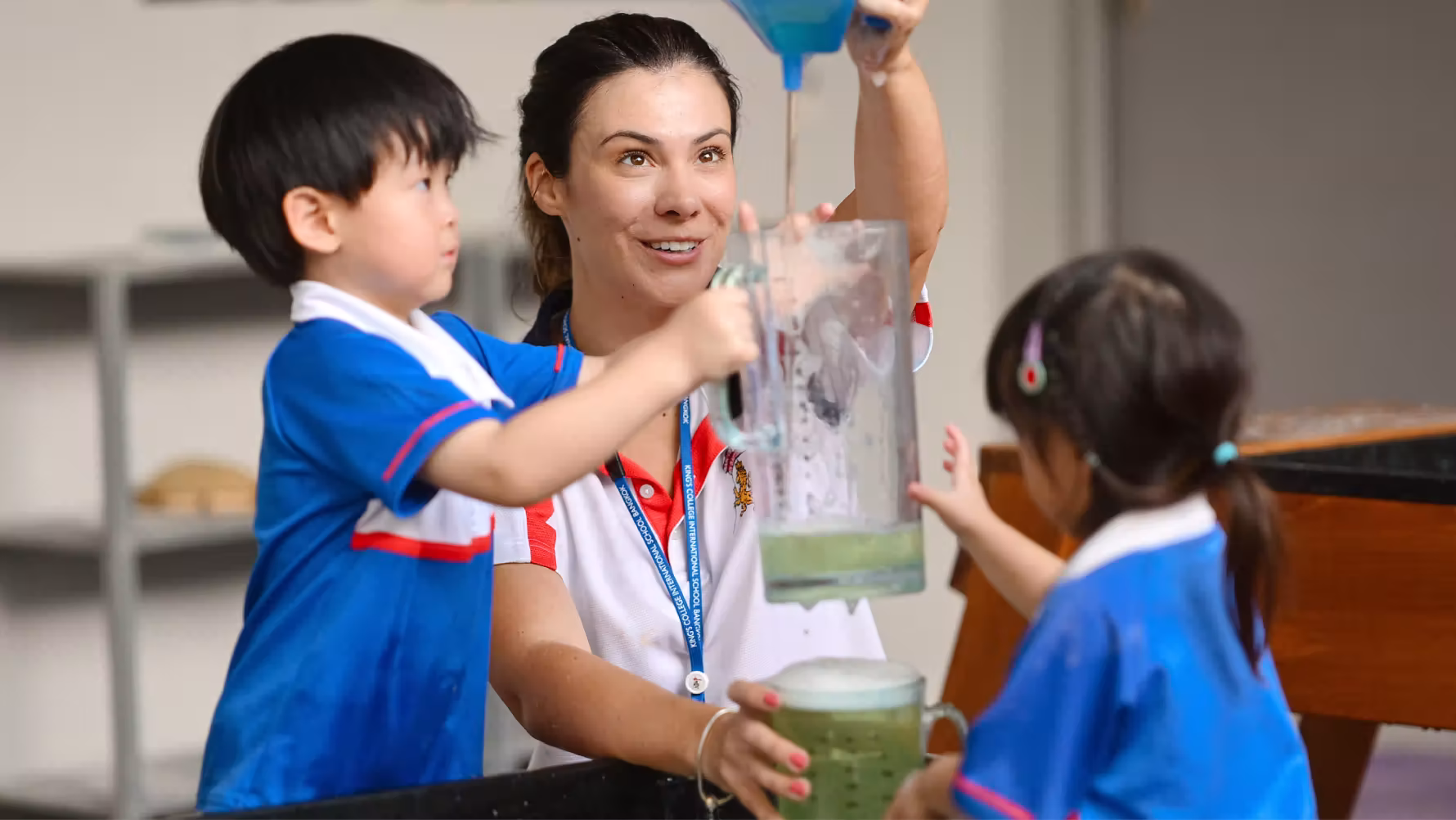 Teacher and two children holding a clear pitcher with green liquid in a classroom setting.