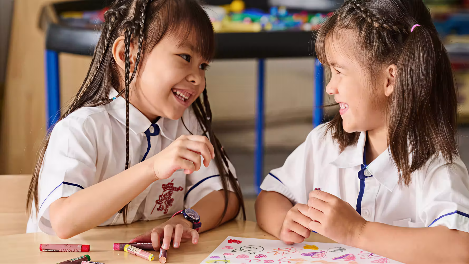 Two young girls in school uniforms smiling and drawing with crayons at a table.