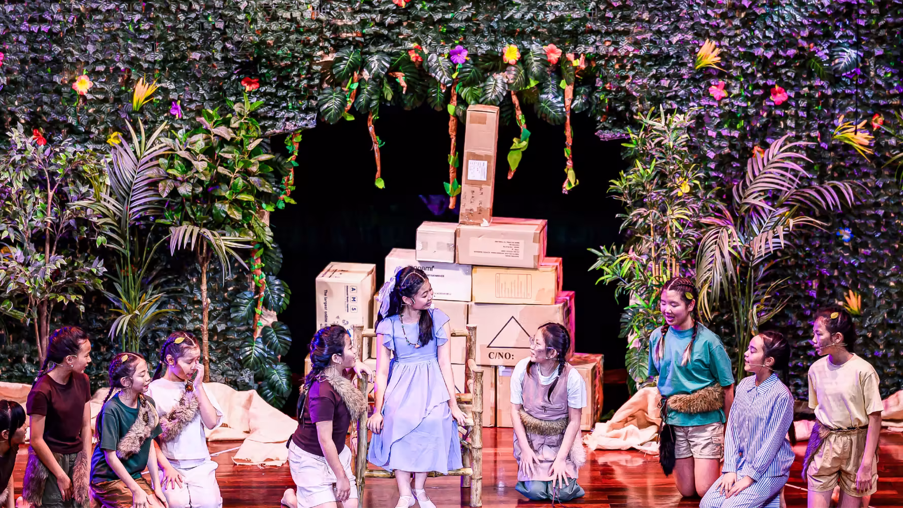 Children in costumes performing on stage with a forest backdrop and cardboard boxes stacked behind them.