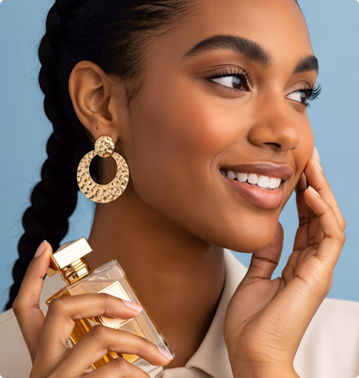 Smiling woman with braided hair holds a gold-capped perfume bottle near her face, wearing large gold hoop earrings.