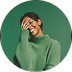 Smiling woman wearing a green sweater covering part of her face with her hand against a green background.