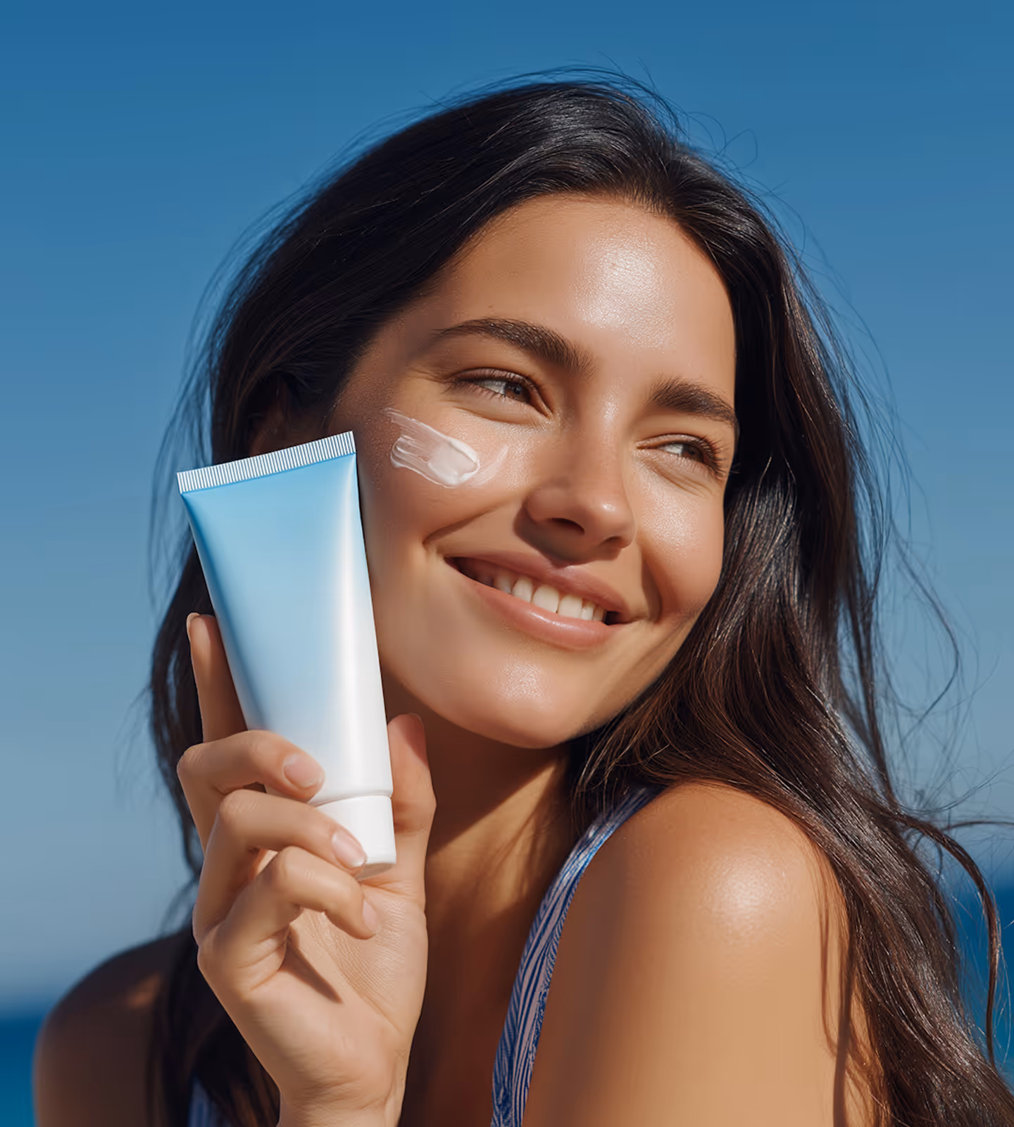 Smiling woman with long dark hair holding a blue and white tube of skincare cream, with a smear of cream on her cheek against a clear blue sky.