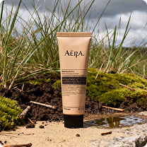A tube of AĒRA skincare product standing upright on sandy ground with moss and grass under a cloudy sky.