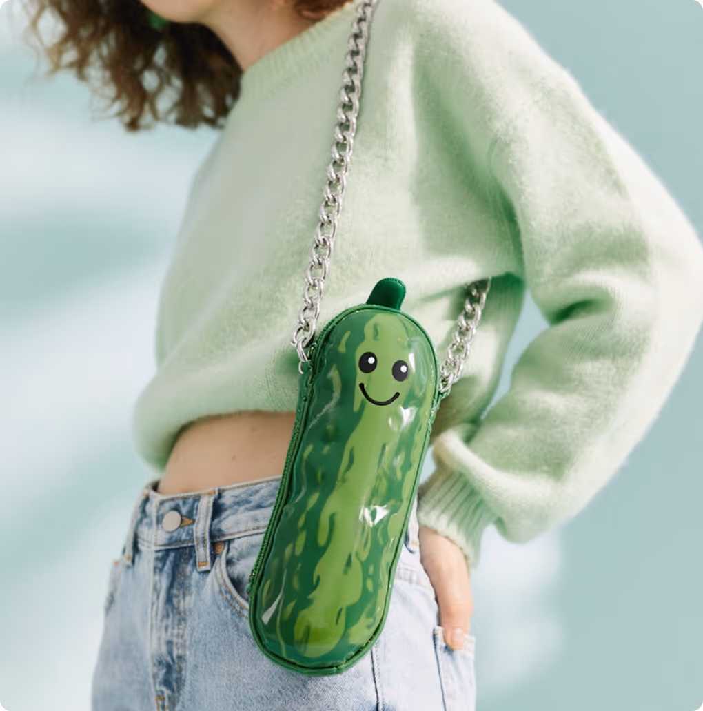 Person wearing light green sweater and blue jeans with a small green pickle-shaped purse with a smiling face and silver chain strap.