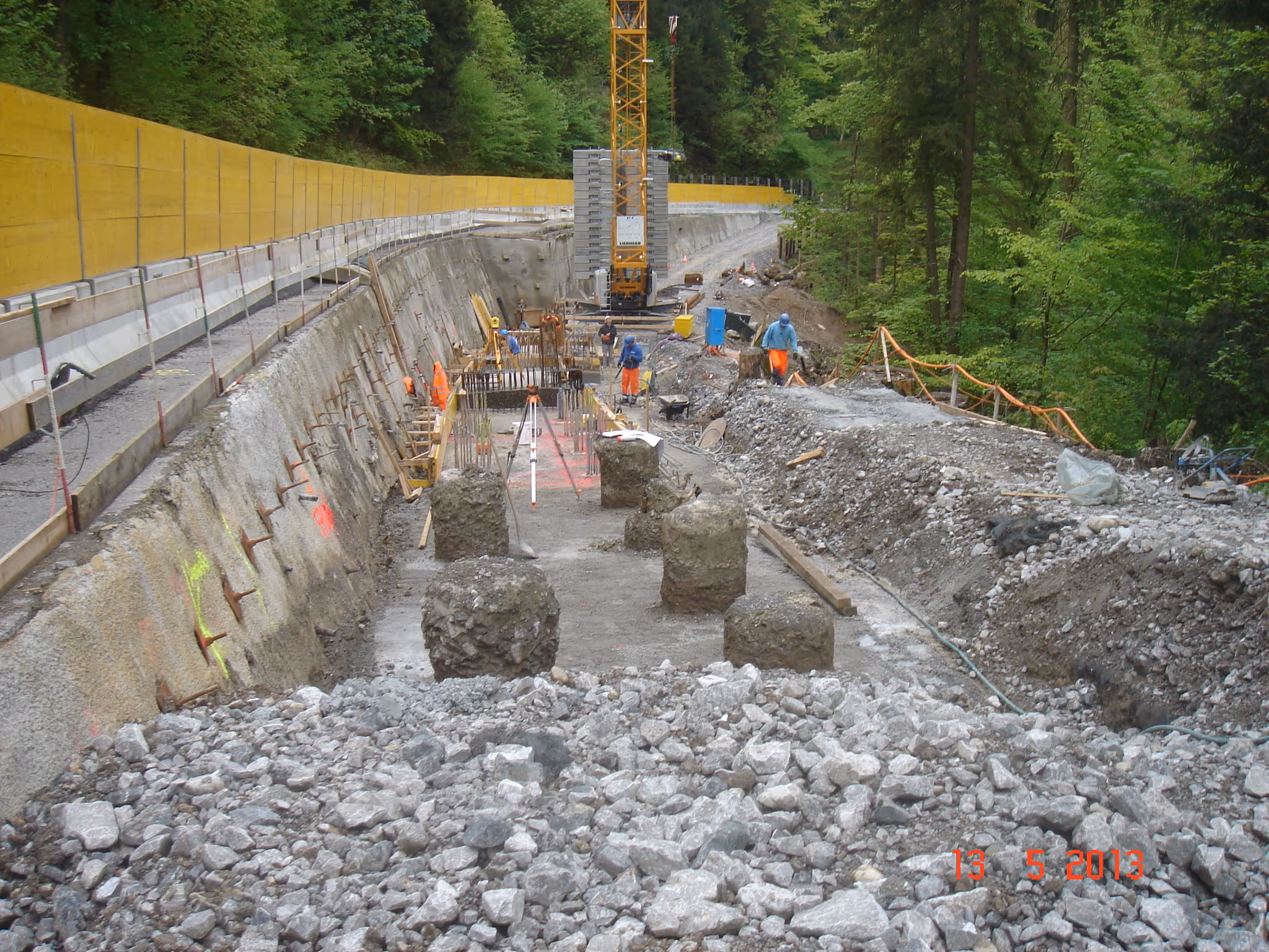 Construction site in a forested area with workers, concrete foundation, and a yellow crane.