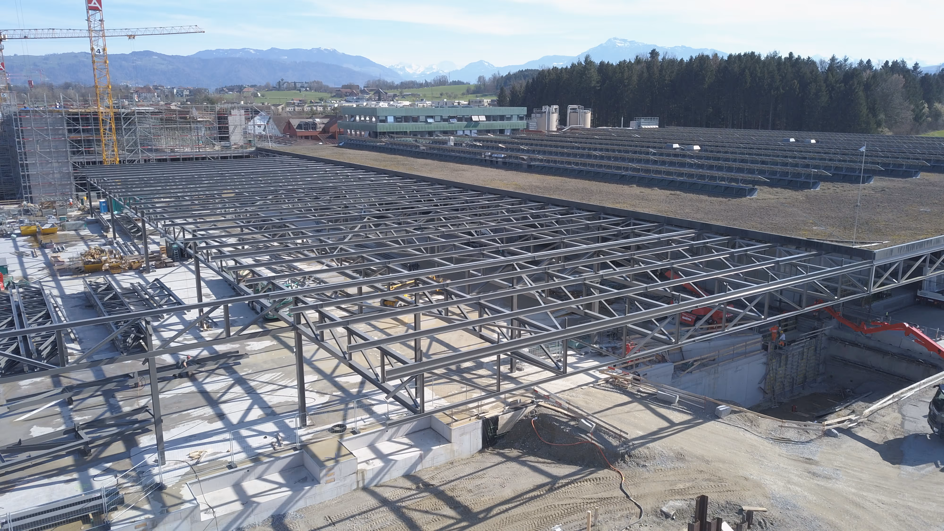 A large construction site with steel framework for a building, surrounded by construction equipment and materials, with mountains and trees in the background.