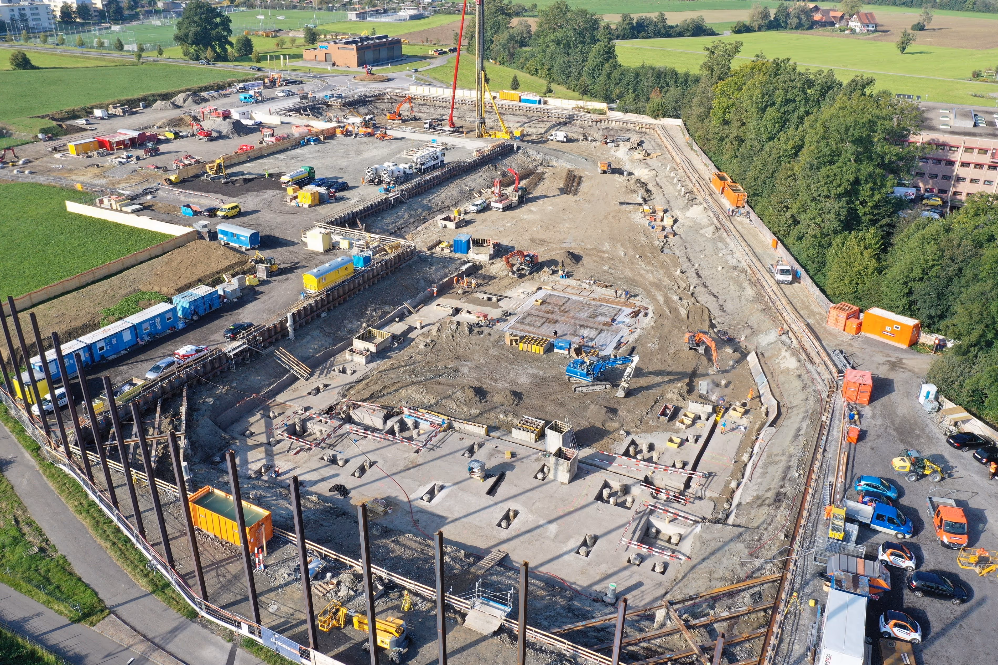 Aerial view of a large construction site with heavy machinery, equipment, and workers, surrounded by green fields and trees.
