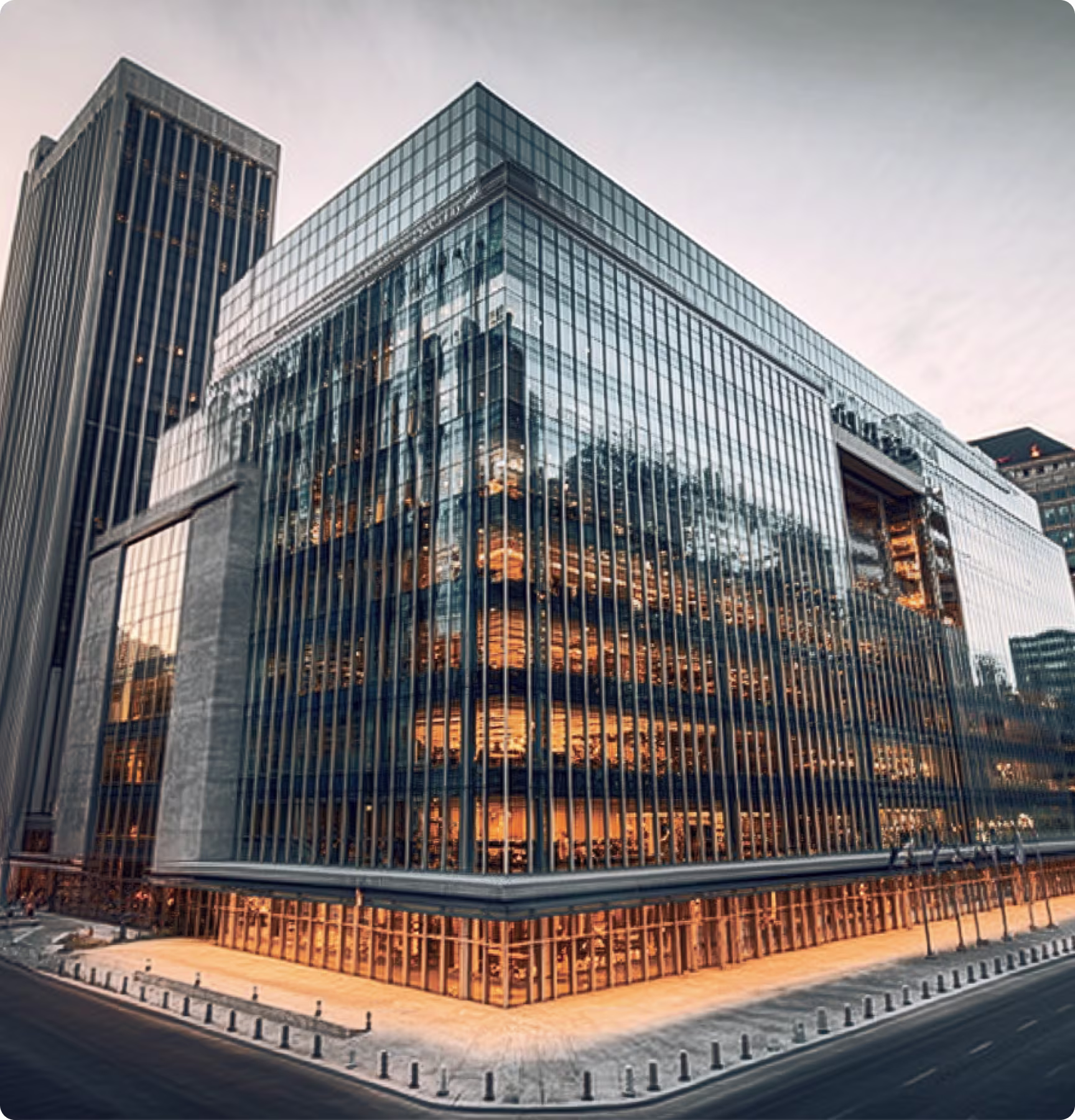 Modern glass office building illuminated warmly at dusk on a city street corner.
