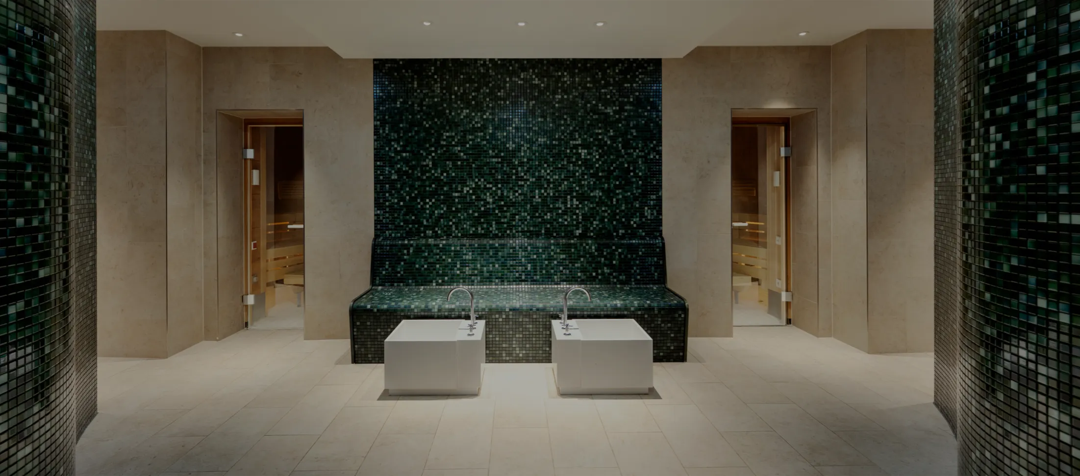 Moderne Sauna-Lounge mit zwei weißen Fußbädern vor einer Sitzbank mit schwarzer Mosaikfliesenwand, flankiert von zwei Türen zu Saunaräumen.