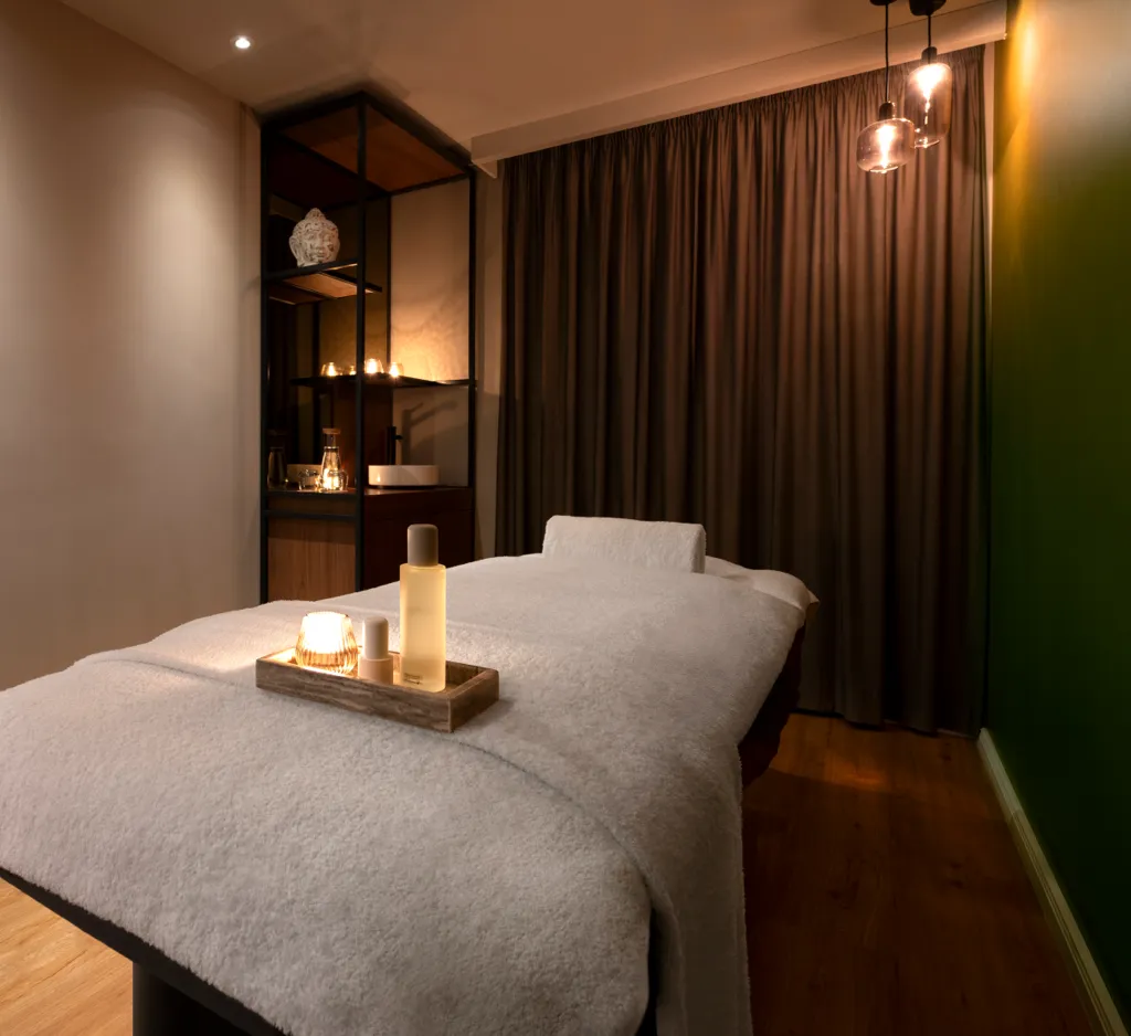 Ein ruhiger Massageraum mit einem mit weißen Decken bedeckten Massagetisch, einer kleinen Holzablage mit Massageöl und einer Kerze, gedämpfte Beleuchtung, Vorhänge und Holzboden.