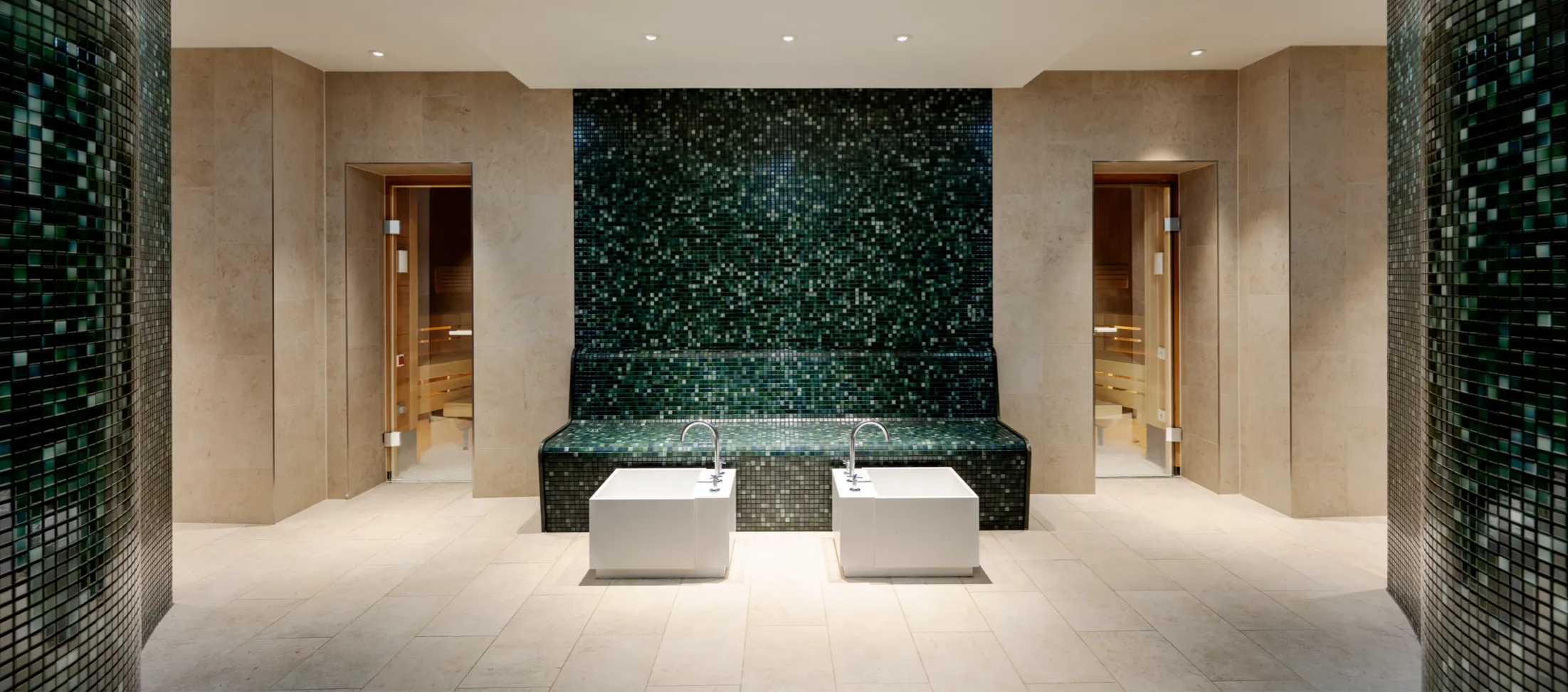 Moderner Spa-Bereich mit hellen Fliesenböden und Wänden aus hellem Stein, zwei offenen Türen mit Saunen dahinter und einem dunklen, glänzenden Mosaik-Sitzbereich in der Mitte, vor dem zwei weiße rechteckige Becken mit Wasserhähnen stehen.