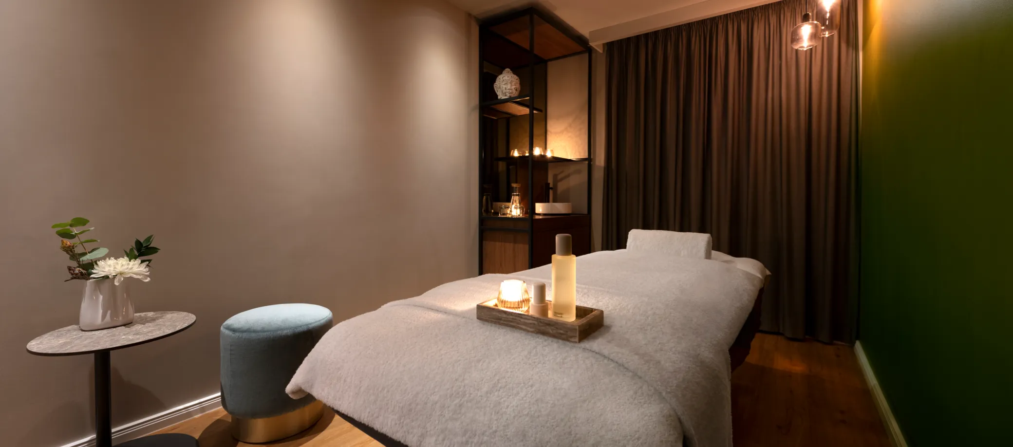 Ruhiger Massageraum mit einem gemütlich bedeckten Massagetisch, einer kleinen Ablage mit Kerze und Flaschen, einem Hocker und einem kleinen Tisch mit Vase und Blumen, umgeben von gedämpften Farben und weichen Vorhängen.