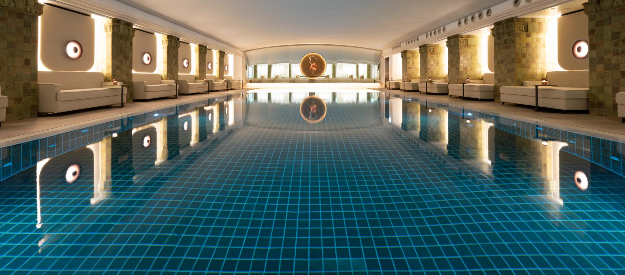 Innenaufnahme eines luxuriösen beheizten Hallenbads mit blau gekacheltem Boden, umgeben von beigen Sesseln, Säulen mit Mosaiksteinverkleidung und runden Wandleuchten, mit einer beleuchteten kreisförmigen Kunstinstallation am Ende.