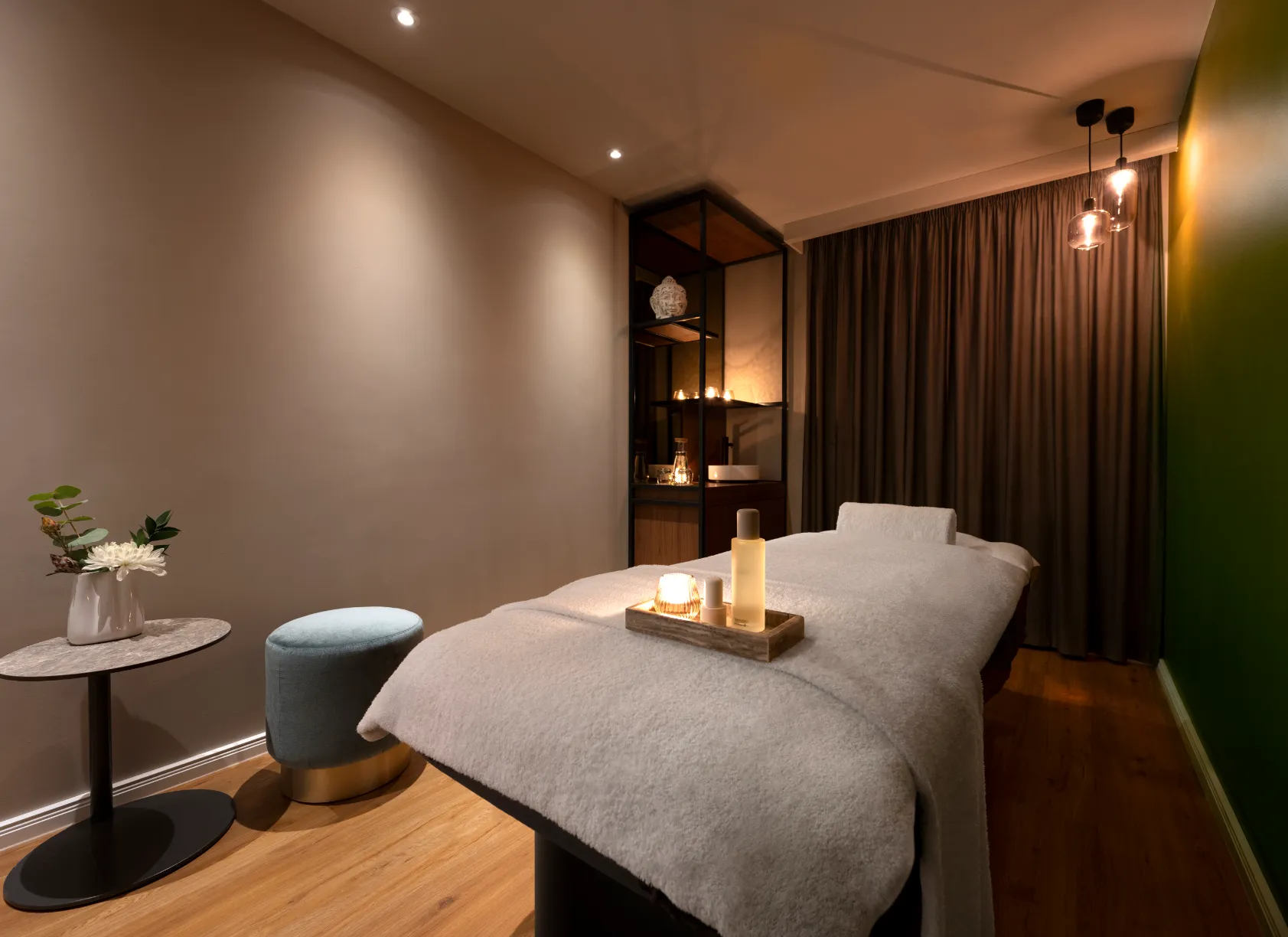 Ruhiger Massageraum mit einem Behandlungstisch, der mit einem weißen Frotteetuch bedeckt ist, einem Holzboden, einem kleinen runden Tisch mit einer Vase und Blumen, samtigem Hocker, Kerzen auf dem Tisch und im Regal, gedämpftem Licht und dunklen Vorhängen.