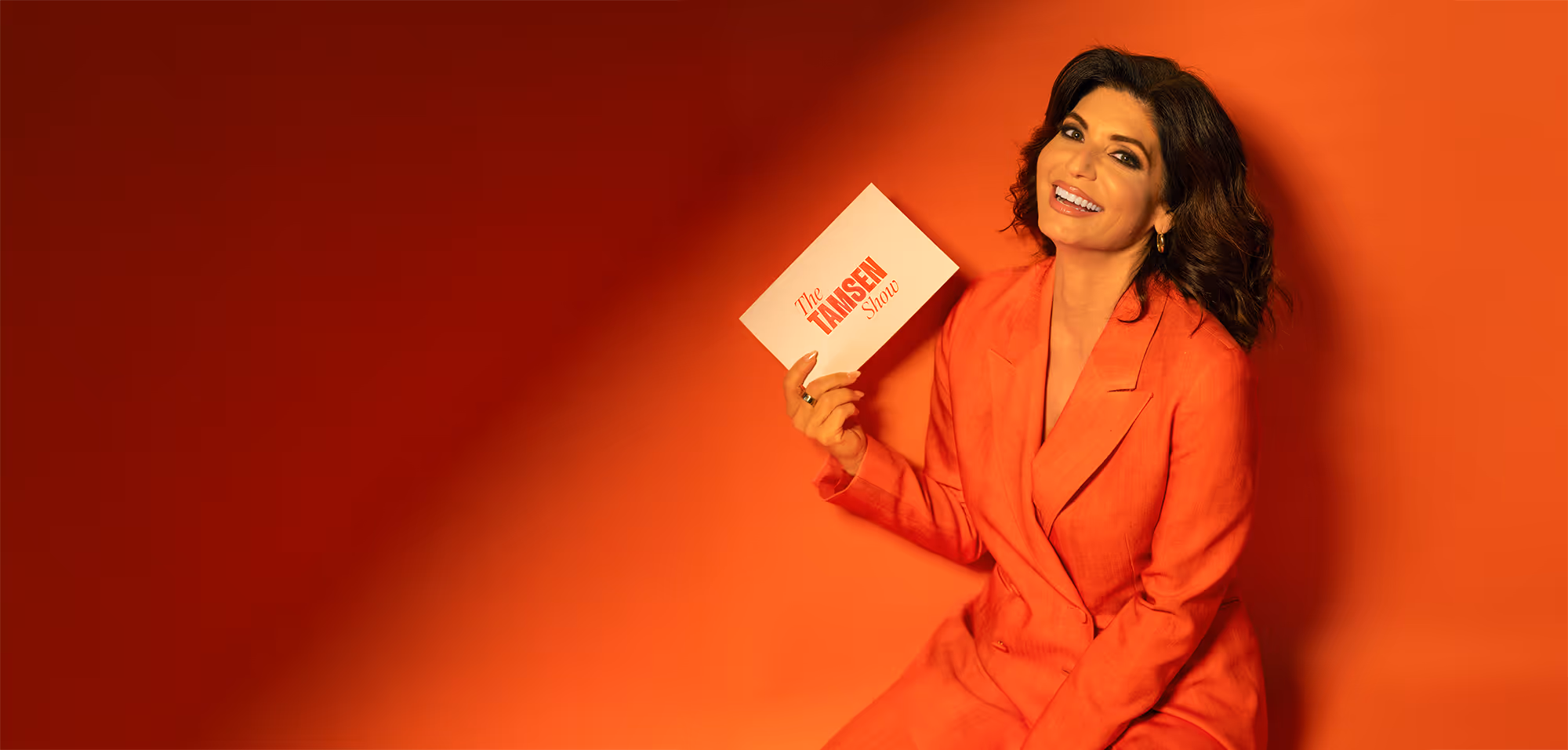Smiling woman in an orange suit sitting against an orange backdrop holding a card reading 'The TAMSEN Show'.