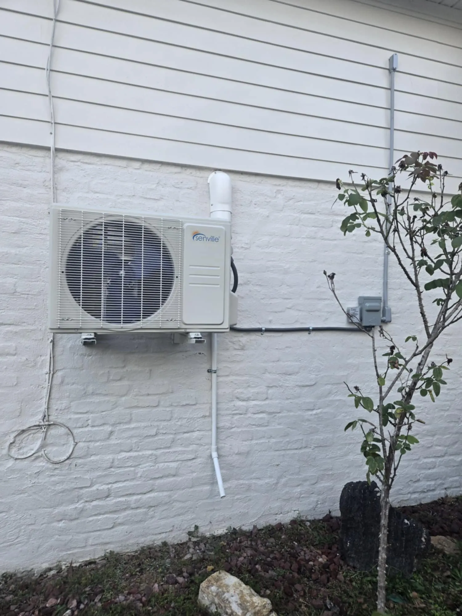 Outdoor air conditioning unit mounted on a white brick wall next to a small tree and rocks.
