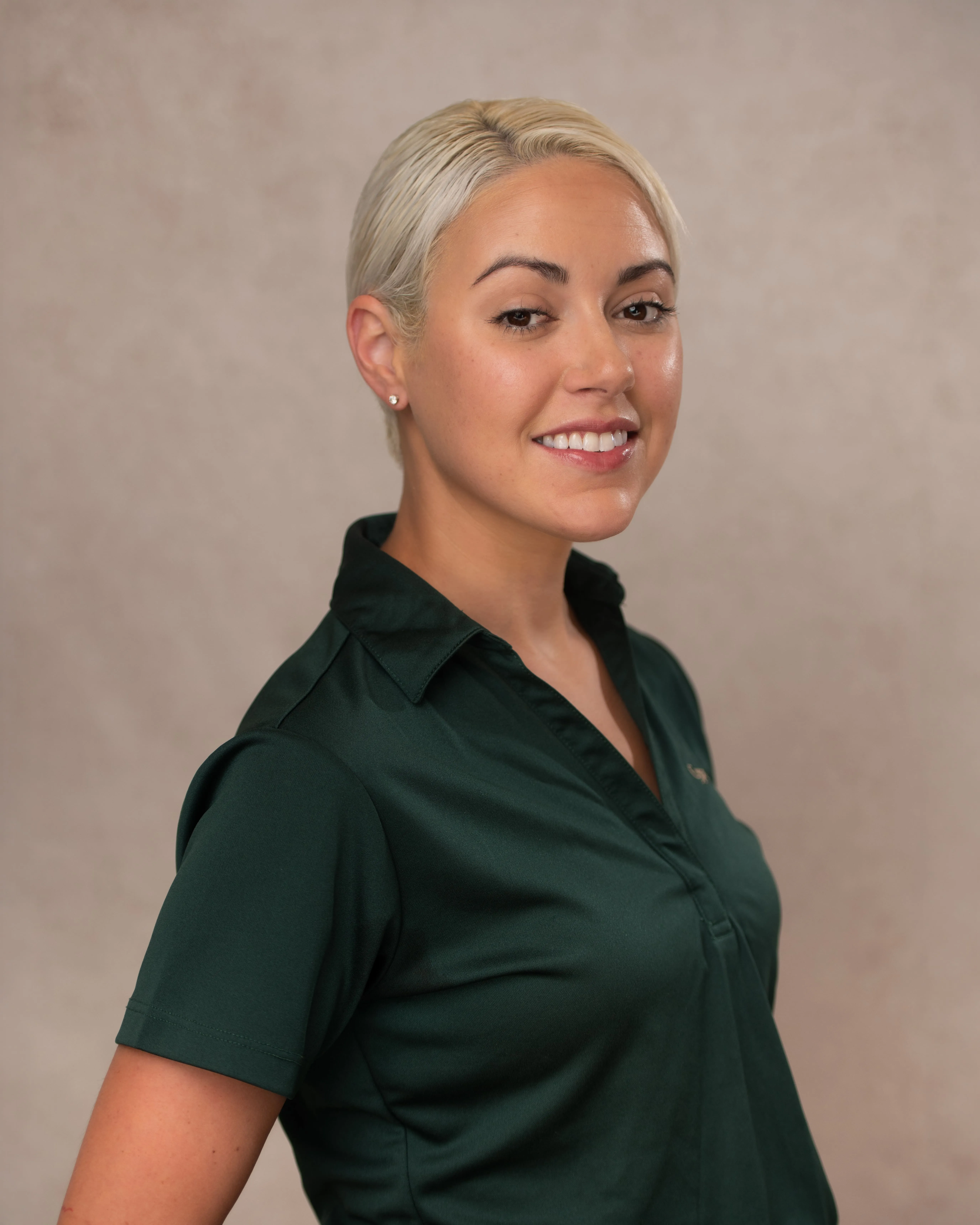 Smiling woman with short blonde hair wearing a dark green collared shirt against a plain background.